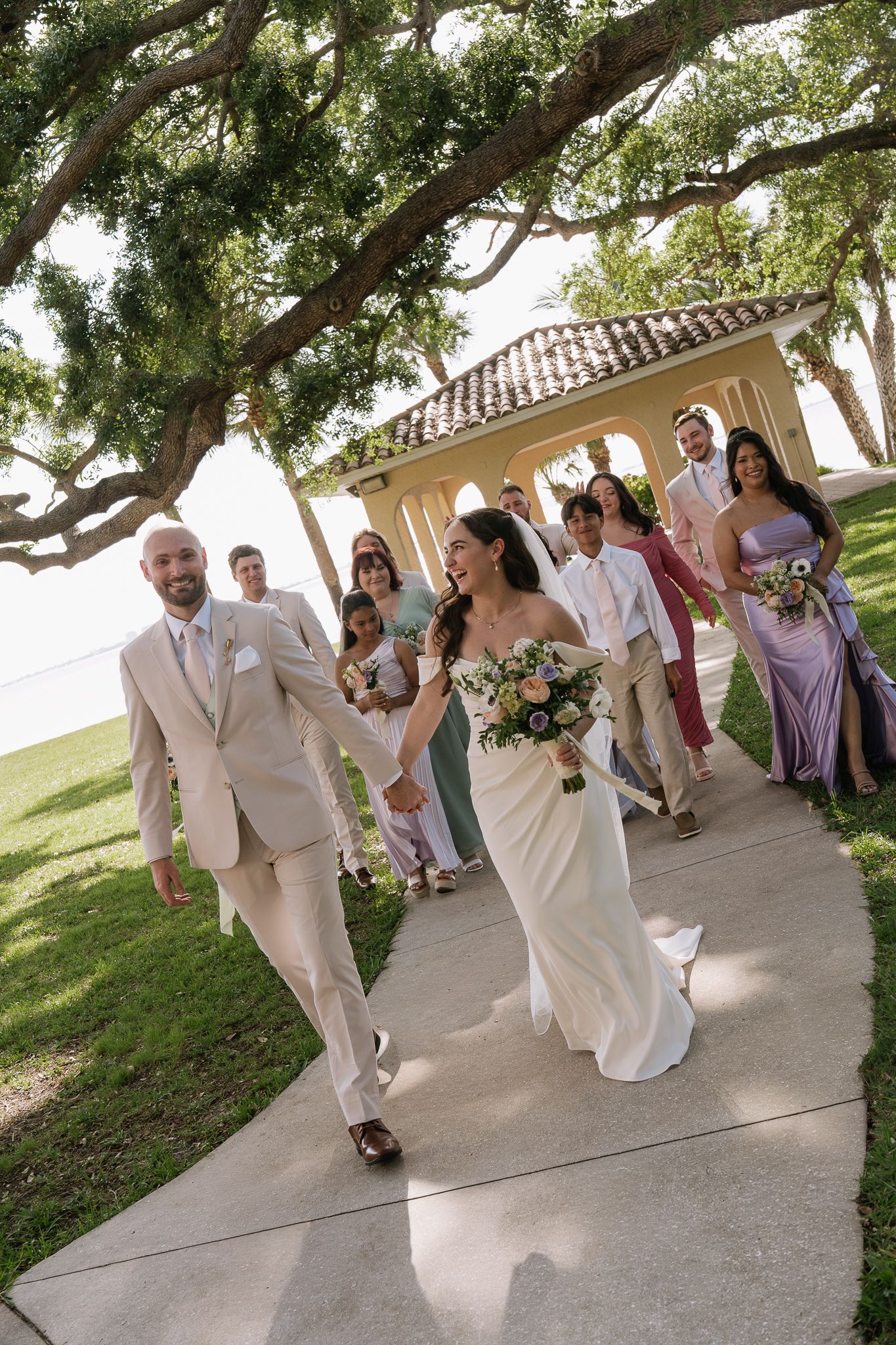 Bride and groom walk hand-in-hand down a sunlit path with wedding party following near a gazebo.