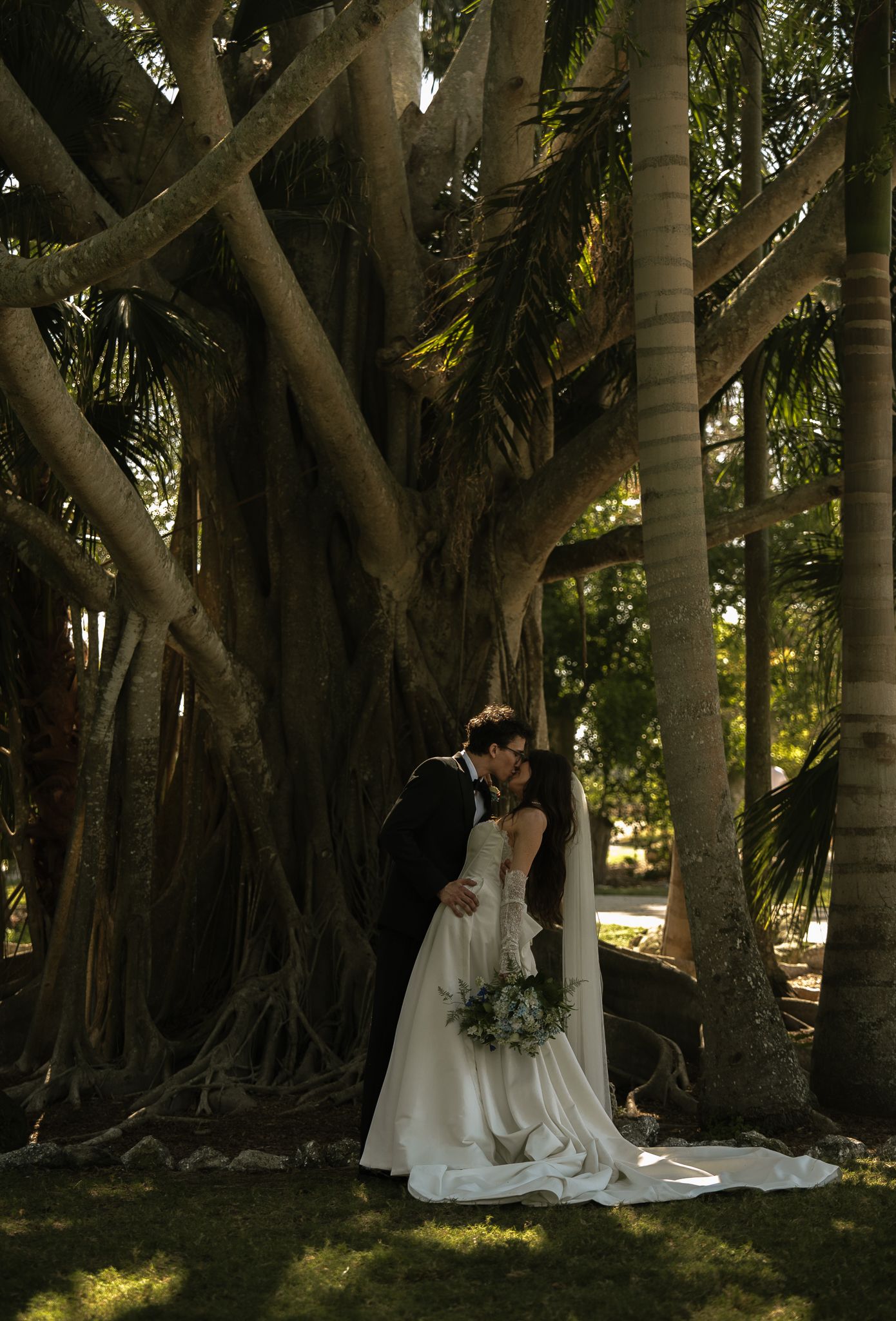 Bride and groom share a romantic kiss beneath a majestic banyan tree, bride holding a bouquet in a flowing white gown.