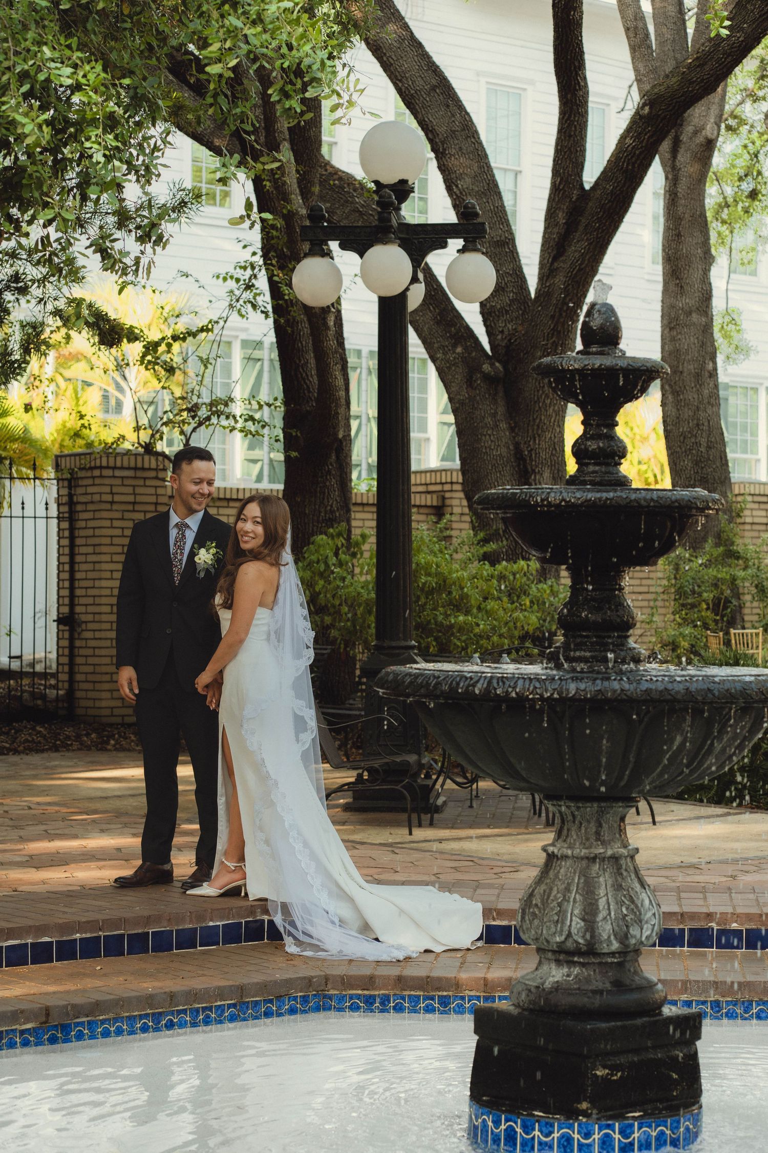 Newlyweds pose beside an ornate black fountain in a sunlit courtyard with lush trees.