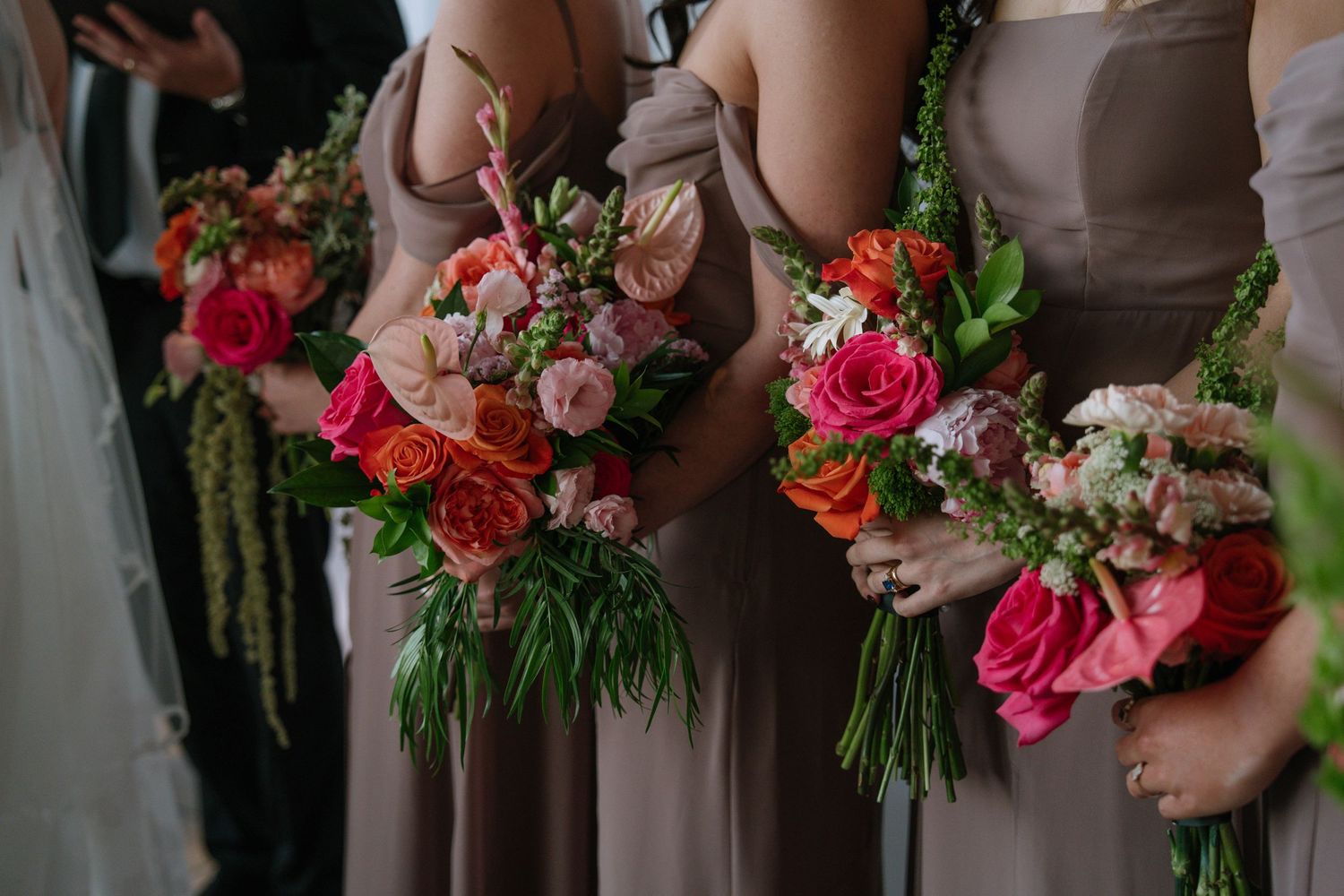 Bridesmaids in taupe dresses hold vibrant bouquets of orange, pink, and red roses with lush greenery.