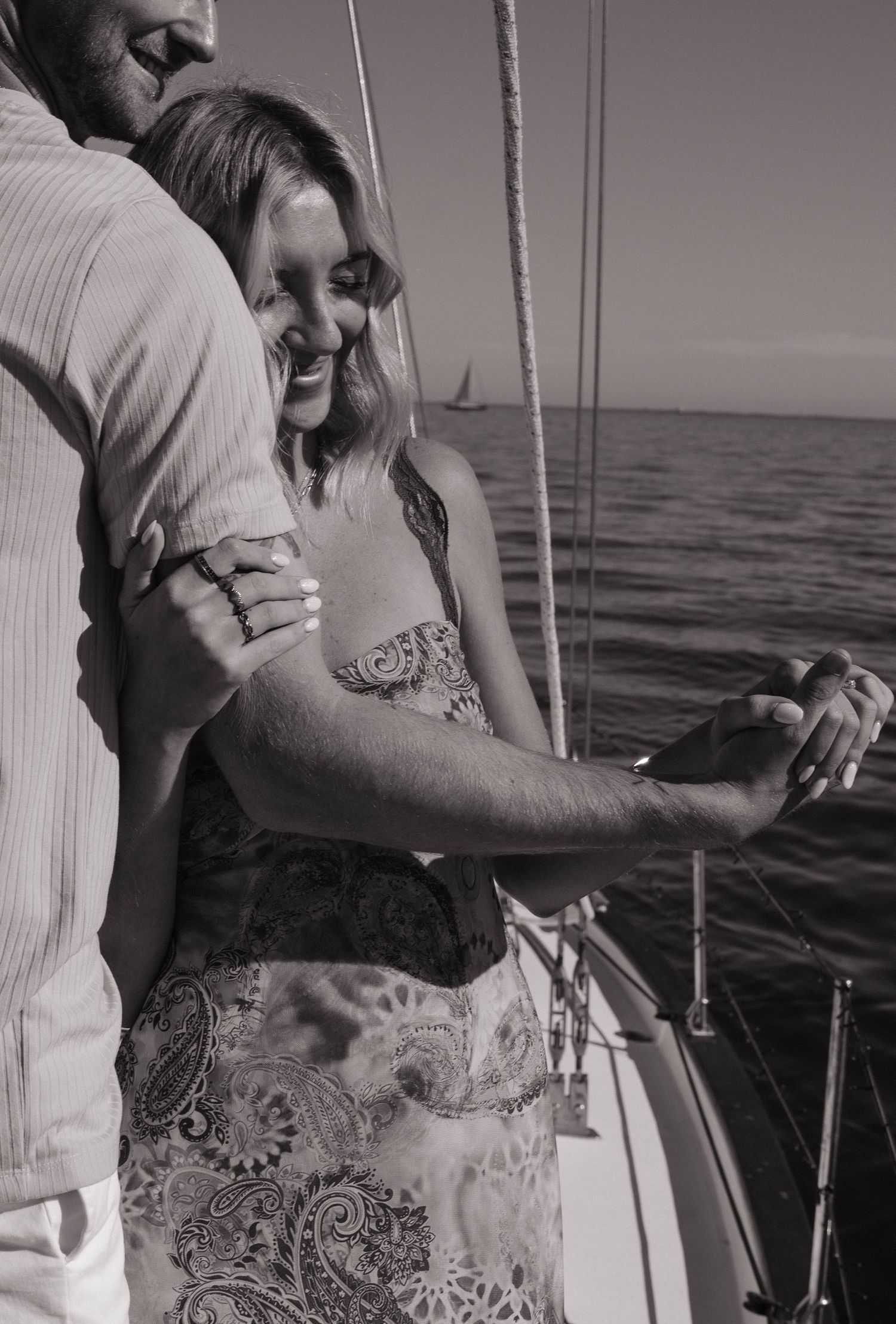 Black and white photo of couple embracing on a sailboat, woman smiling with engagement ring.