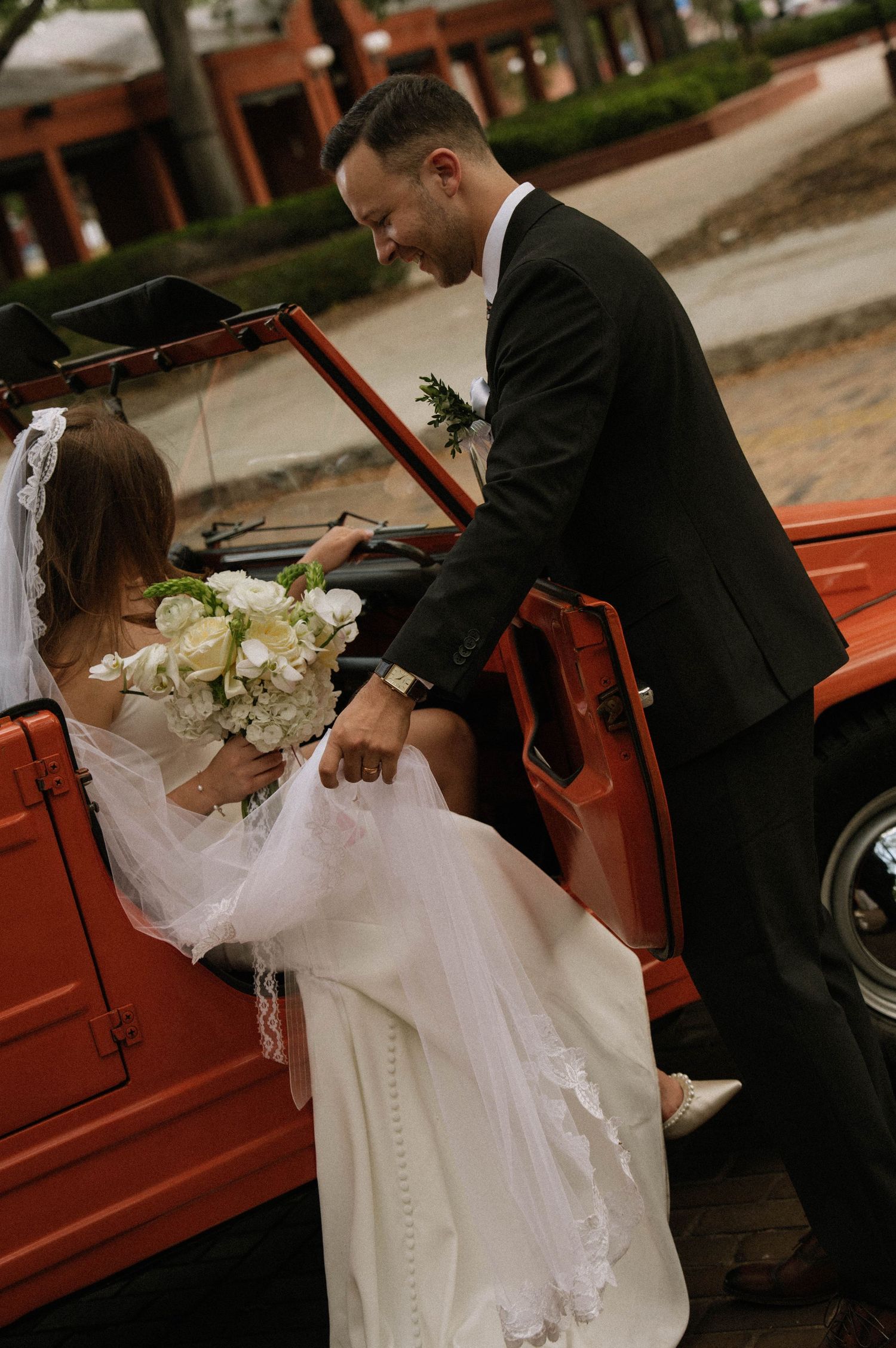 Groom helps bride in white gown with bouquet into a vintage red convertible car after wedding ceremony.