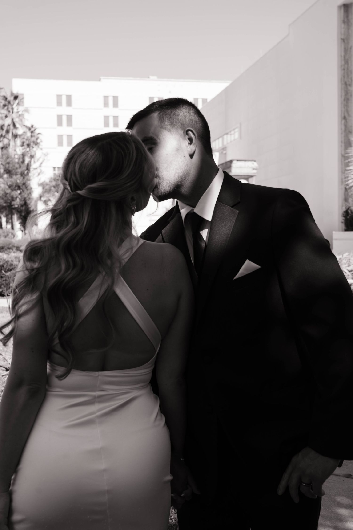 Black and white photo of couple kissing outdoors, woman in elegant backless dress, man in dark suit.