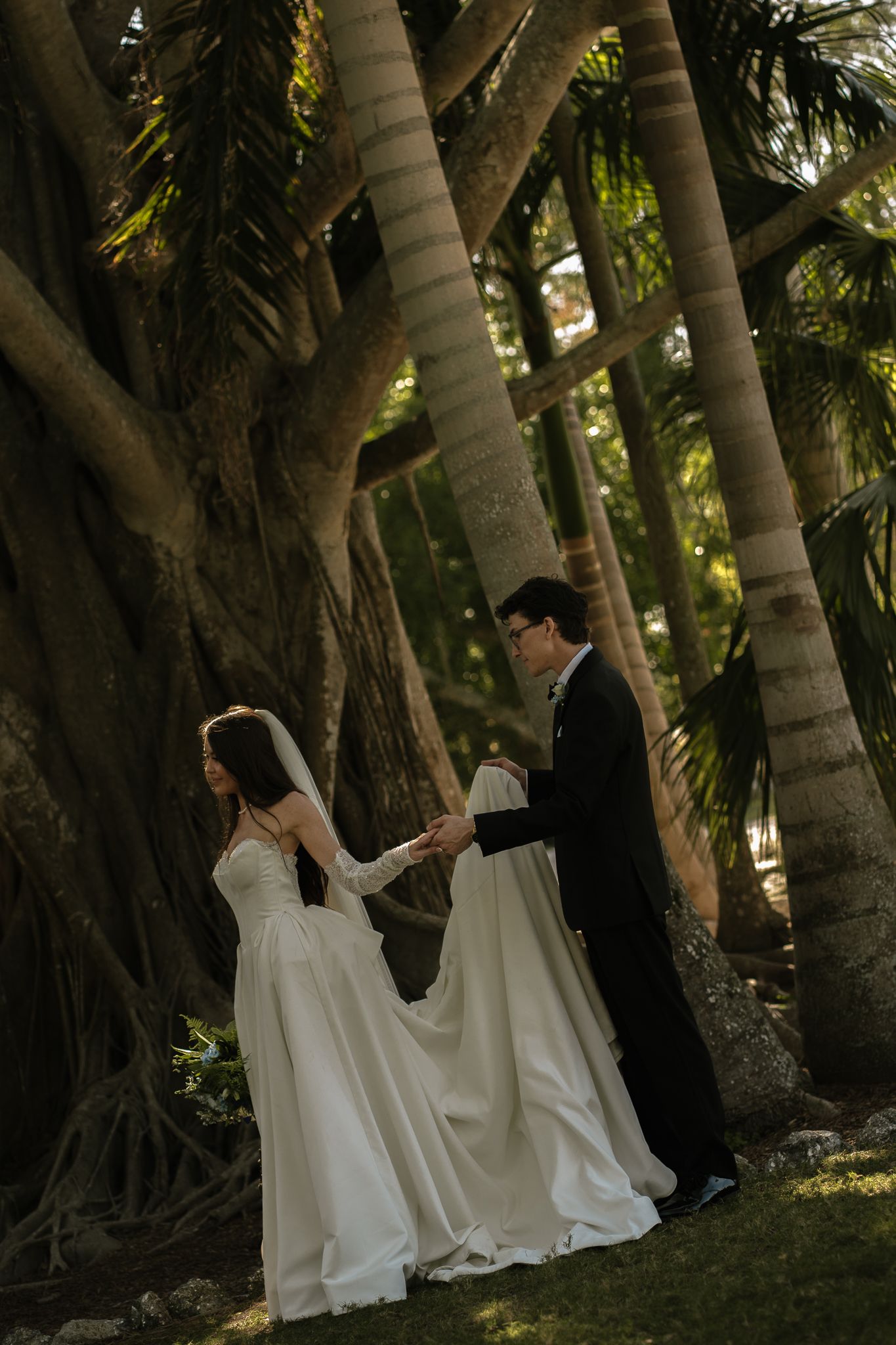 Bride and groom walk hand in hand among towering palm trees in lush tropical garden setting.