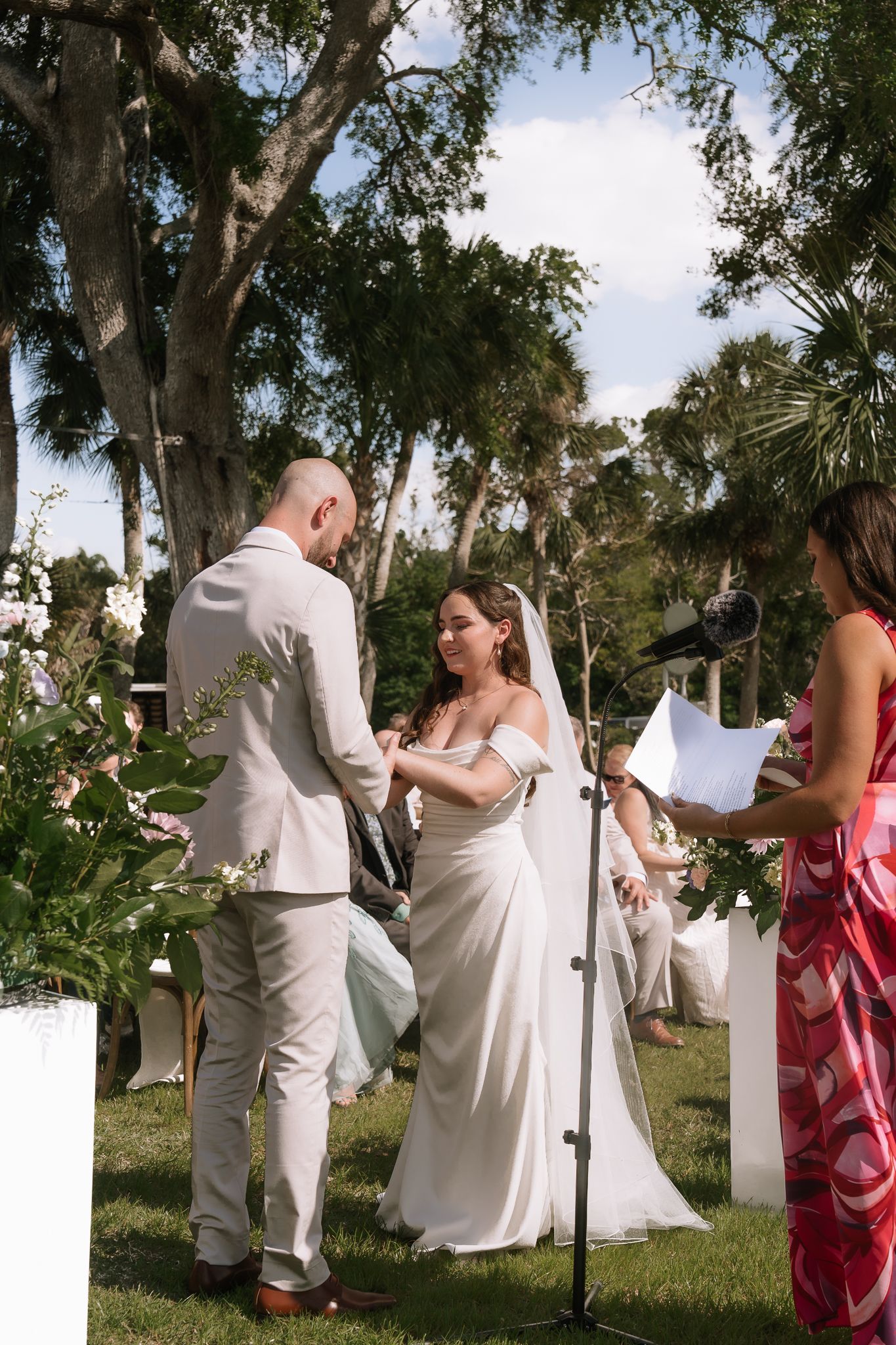 Bride and groom exchange vows at an outdoor garden ceremony with lush trees and floral arrangements.