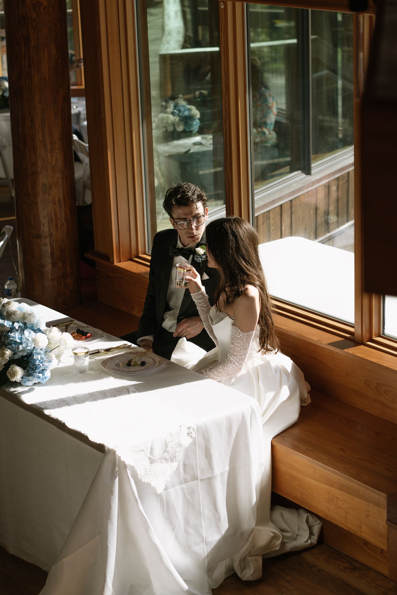 Bride and groom sharing an intimate moment at sweetheart table by sunlit window during wedding reception.
