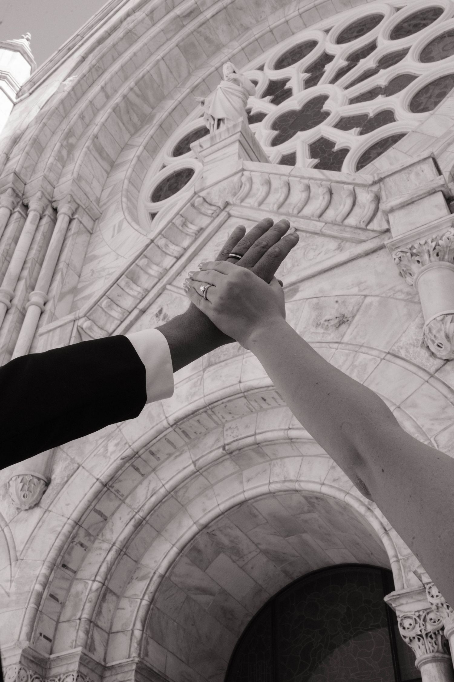Black and white photo of a bride and groom raising joined hands before an ornate cathedral facade.