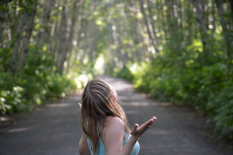 A young girl with long hair stands on a tree-lined path reaching her hand outward.