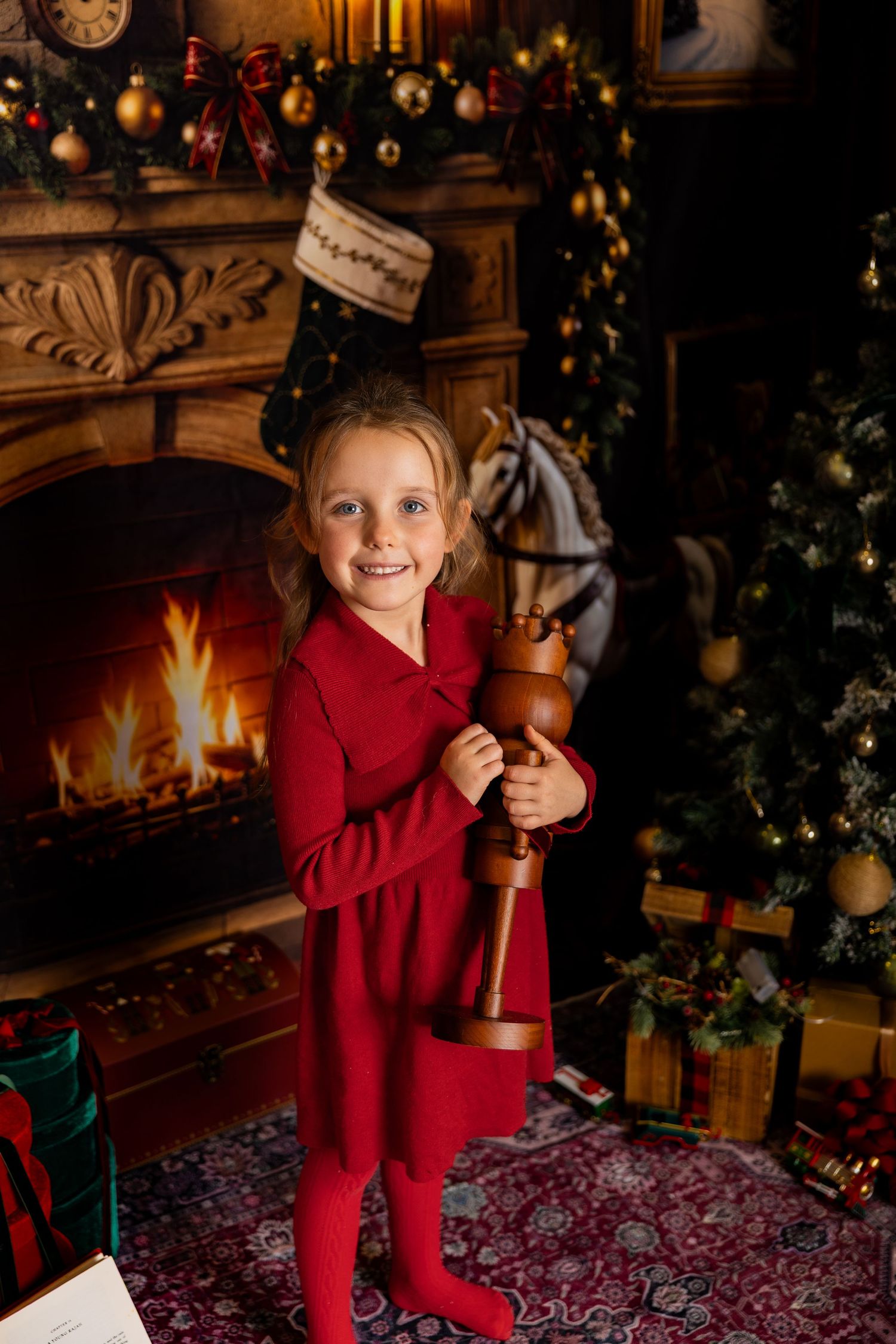 Smiling girl in a red dress holds a wooden nutcracker toy by a glowing fireplace and a lit Christmas tree with presents.