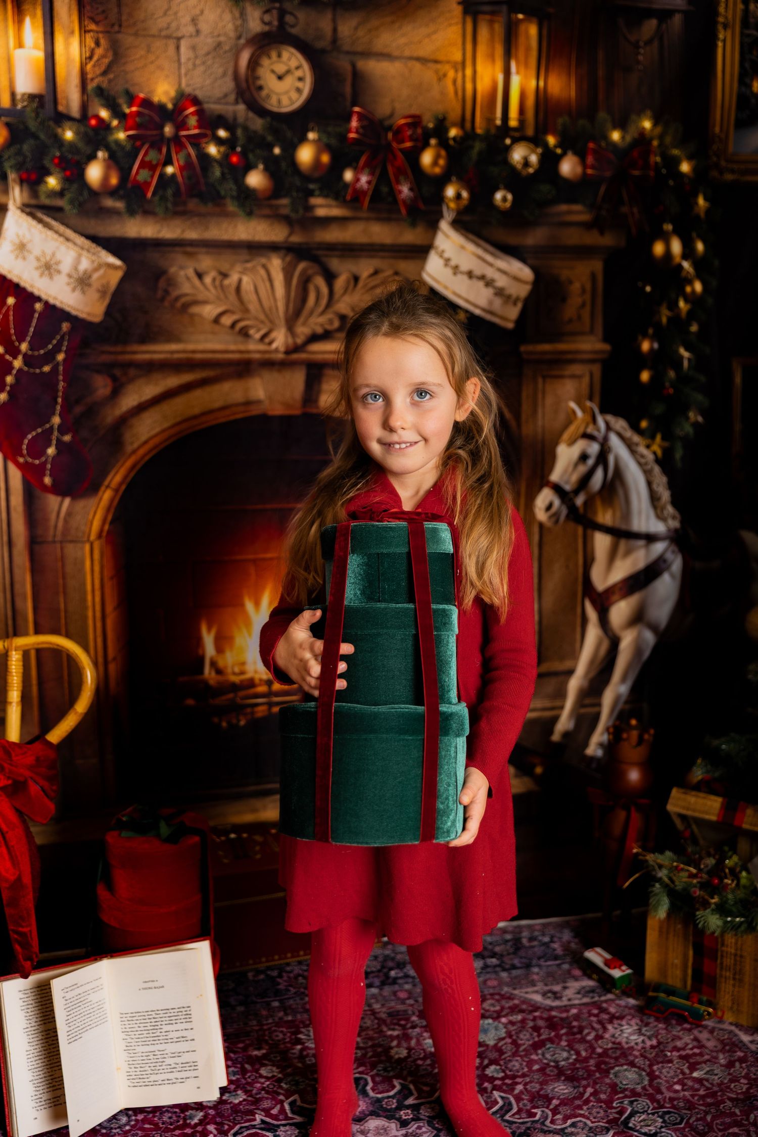 Smiling girl in red holds a stack of green velvet gift boxes before a warm fireplace with Christmas stockings and rocking horse.