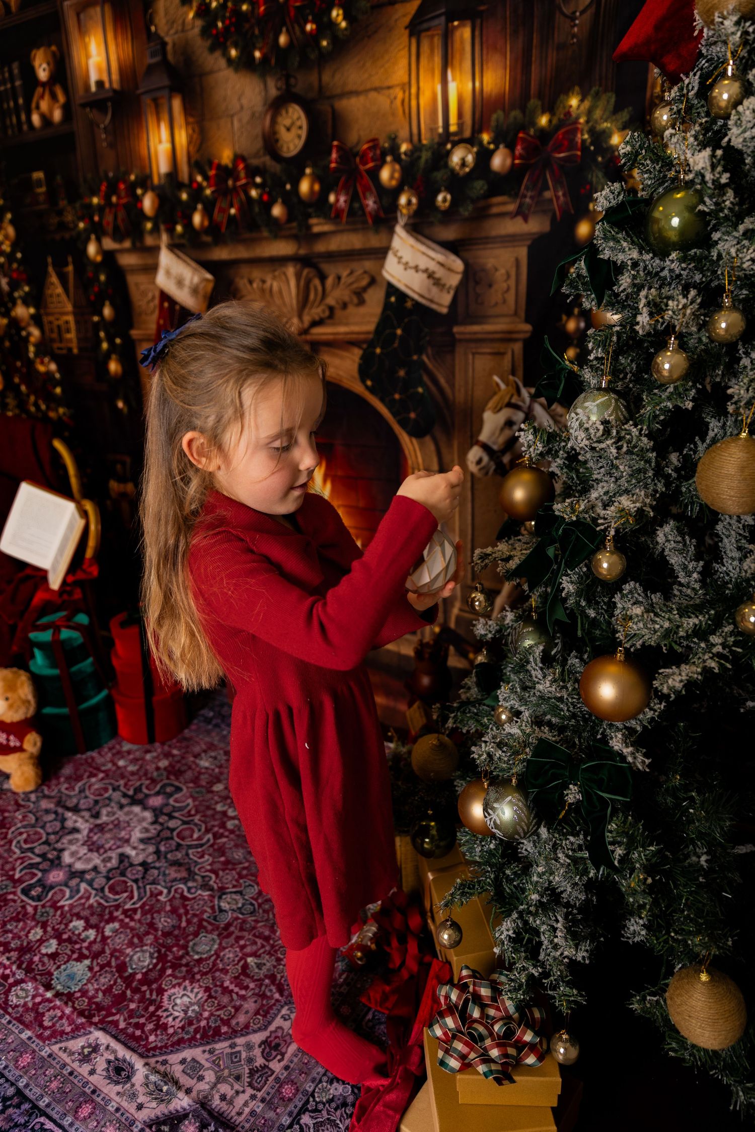 Girl in a red dress decorates a frosted Christmas tree with gold ornaments beside a cozy fireplace and hanging stockings.