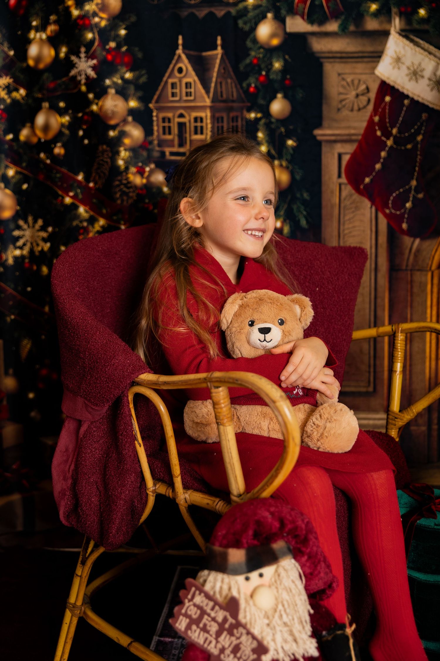Smiling girl in red holds a teddy bear in a wicker rocking chair beside a decorated Christmas tree and stocking.