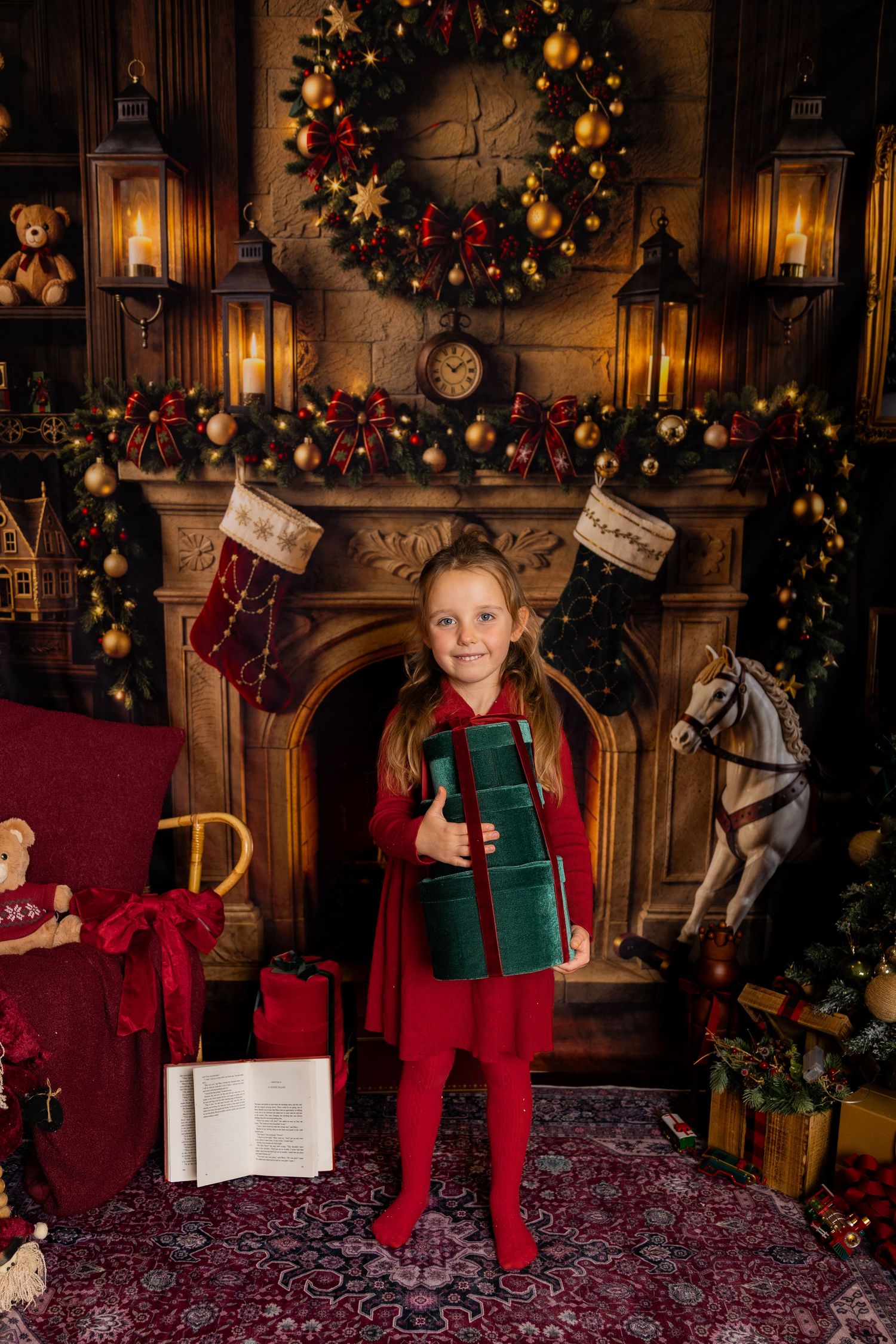 Girl in a red dress holds a green gift box in front of a decorated mantel with stockings, wreath, and rocking horse.
