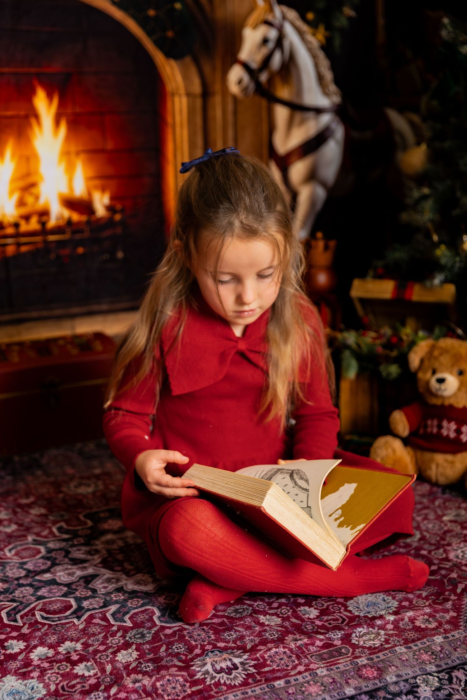 Girl in red sits on a patterned rug reading a large book by a crackling fireplace with a rocking horse and teddy bear.