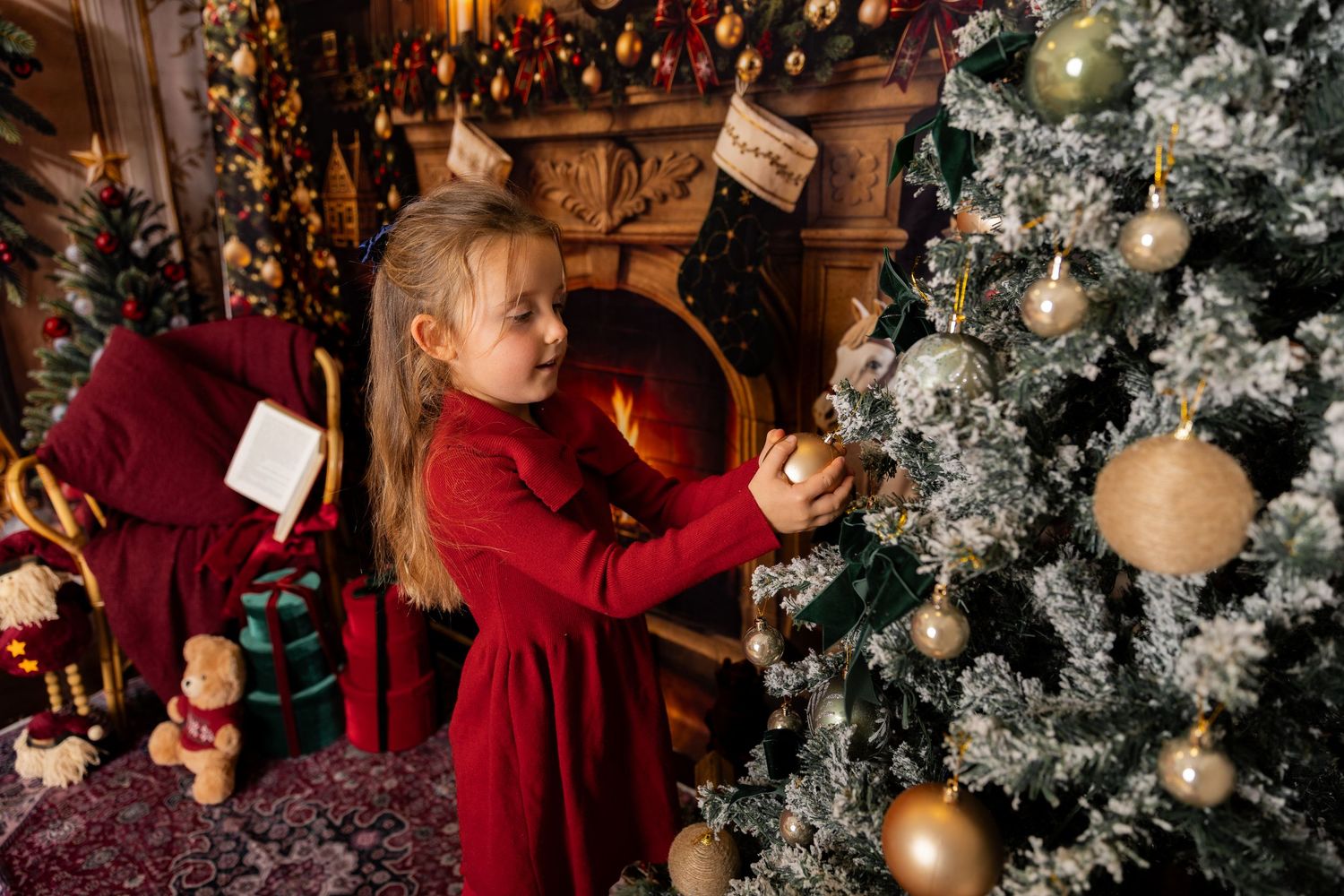 Little girl in red hangs a gold bauble on a flocked Christmas tree in a festive room with fireplace and teddy bear.