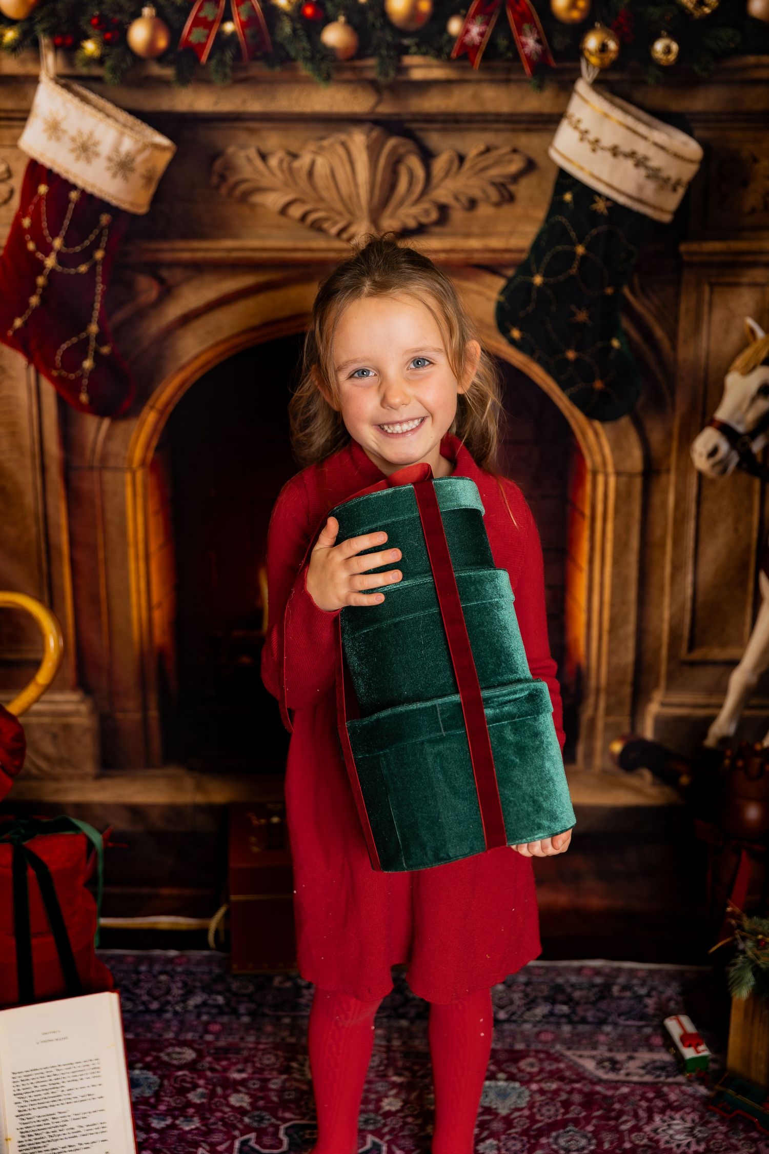 Happy girl in a red dress cradles stacked green velvet gift boxes in front of a Christmas fireplace with hanging stockings.