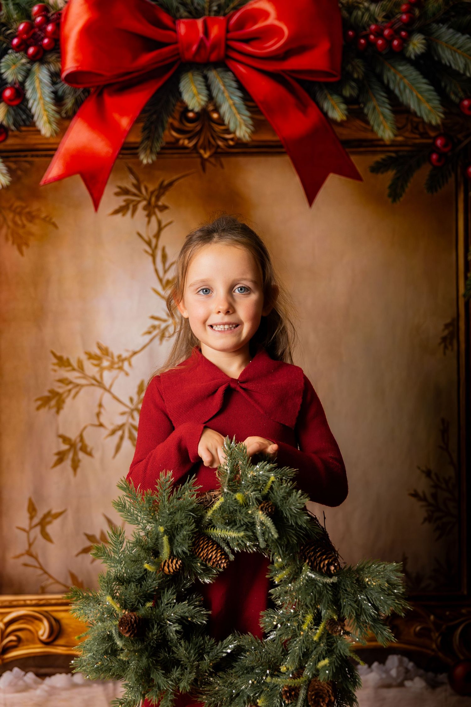 Girl in a red dress holds a pine wreath with pinecones beneath a big red bow in a festive holiday portrait.