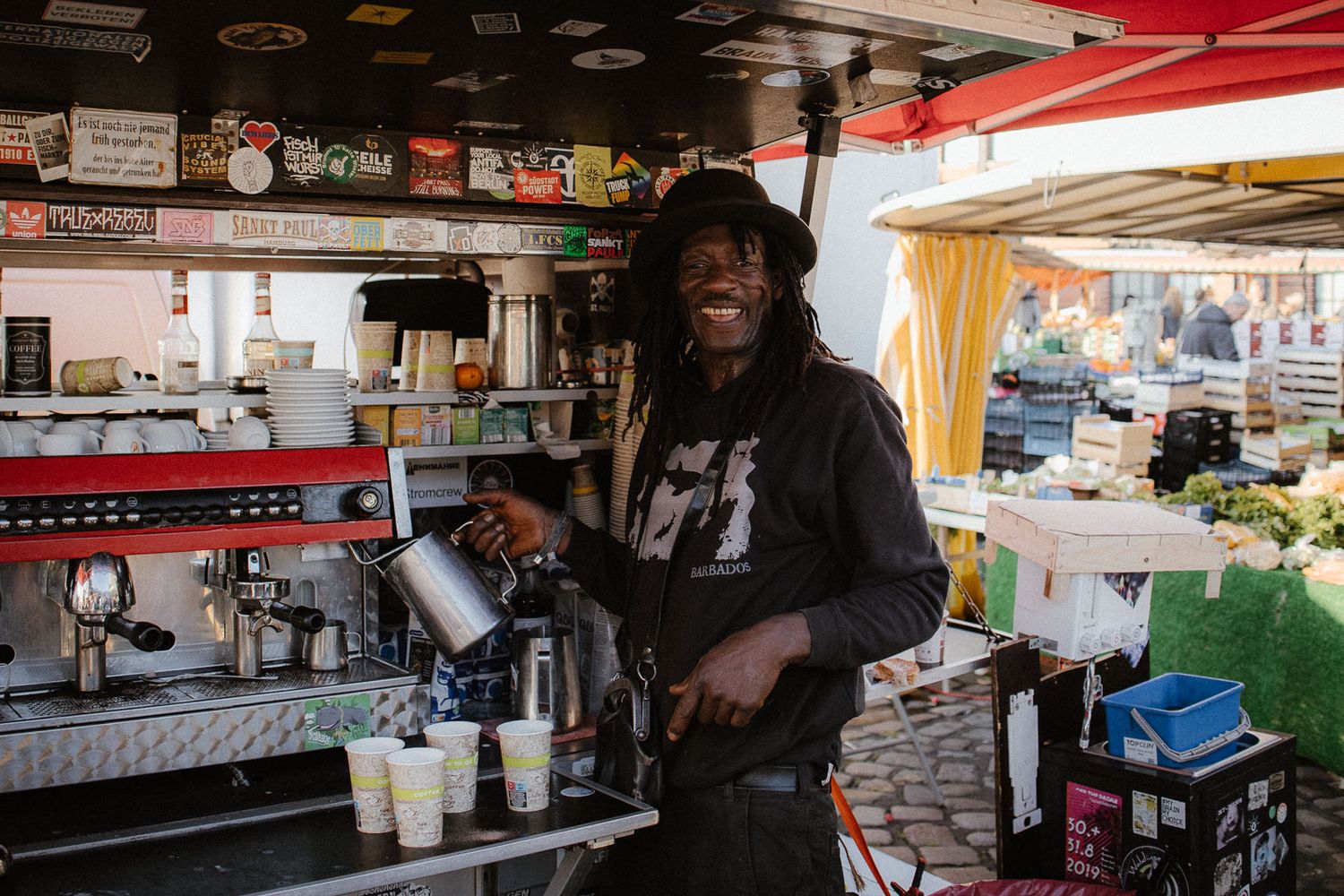 A smiling barista pours milk at an outdoor espresso coffee stand at a vibrant street market.