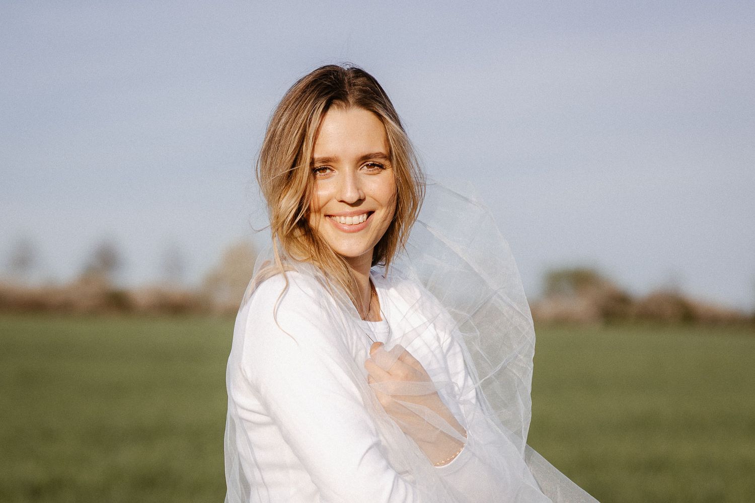 Smiling bride in white dress holding veil in green field on sunny day.