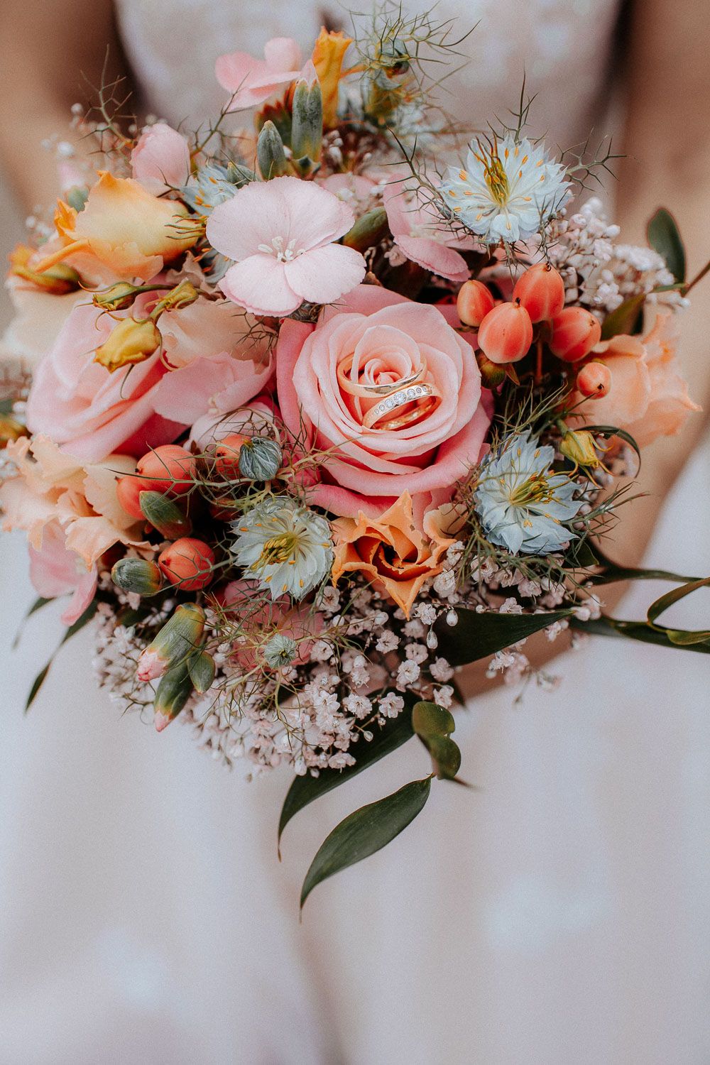 Bride holding a lush wedding bouquet with pink roses, orange flowers, and baby's breath featuring gold rings.