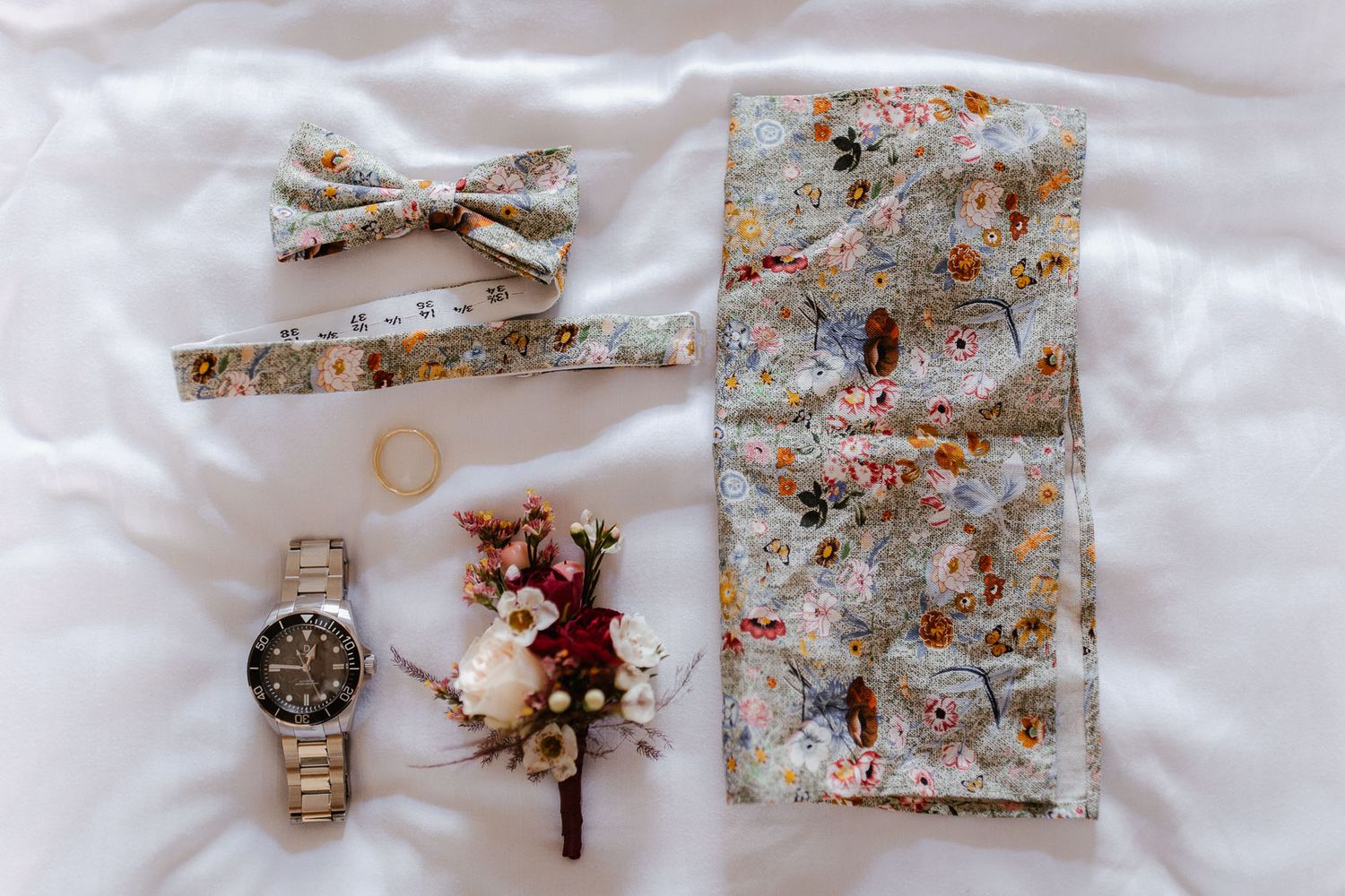 Floral bow tie, pocket square, boutonniere, watch, and gold ring arranged on white fabric for a wedding flat lay.