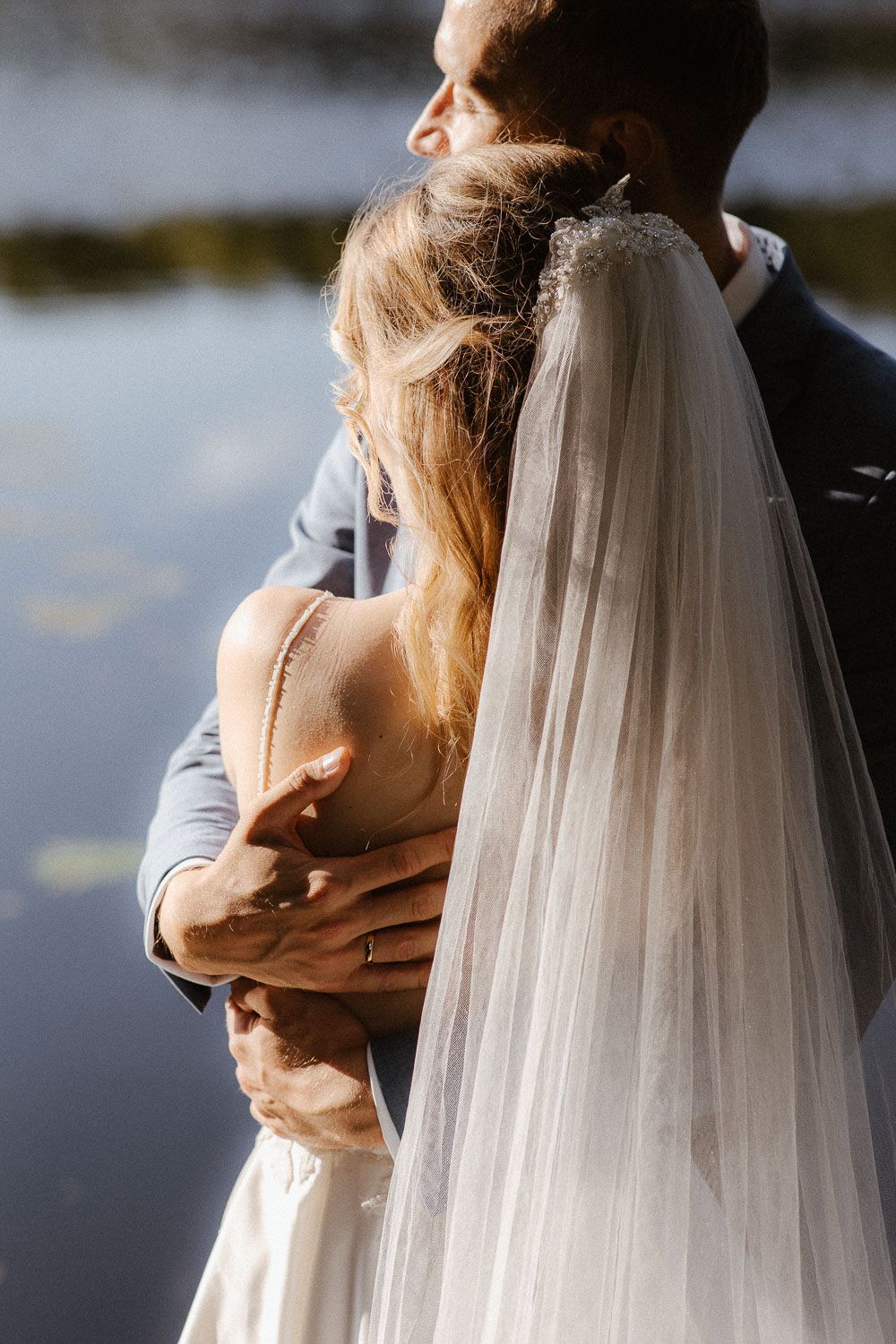 Groom embracing bride from behind near a reflective lake, showcasing her flowing veil in golden sunlight.