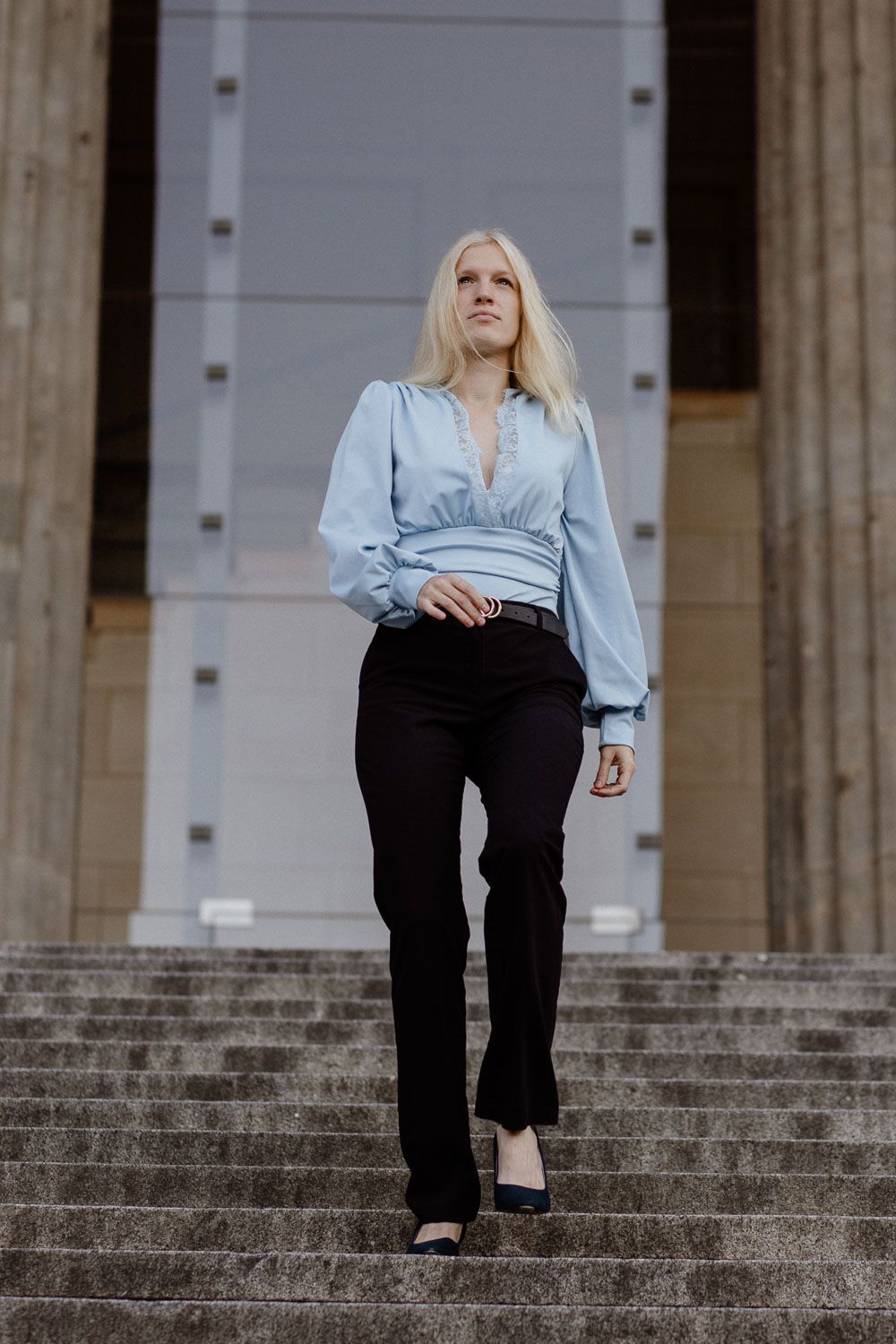 Blonde woman in blue satin blouse and black pants walking down stone steps of a classical building.