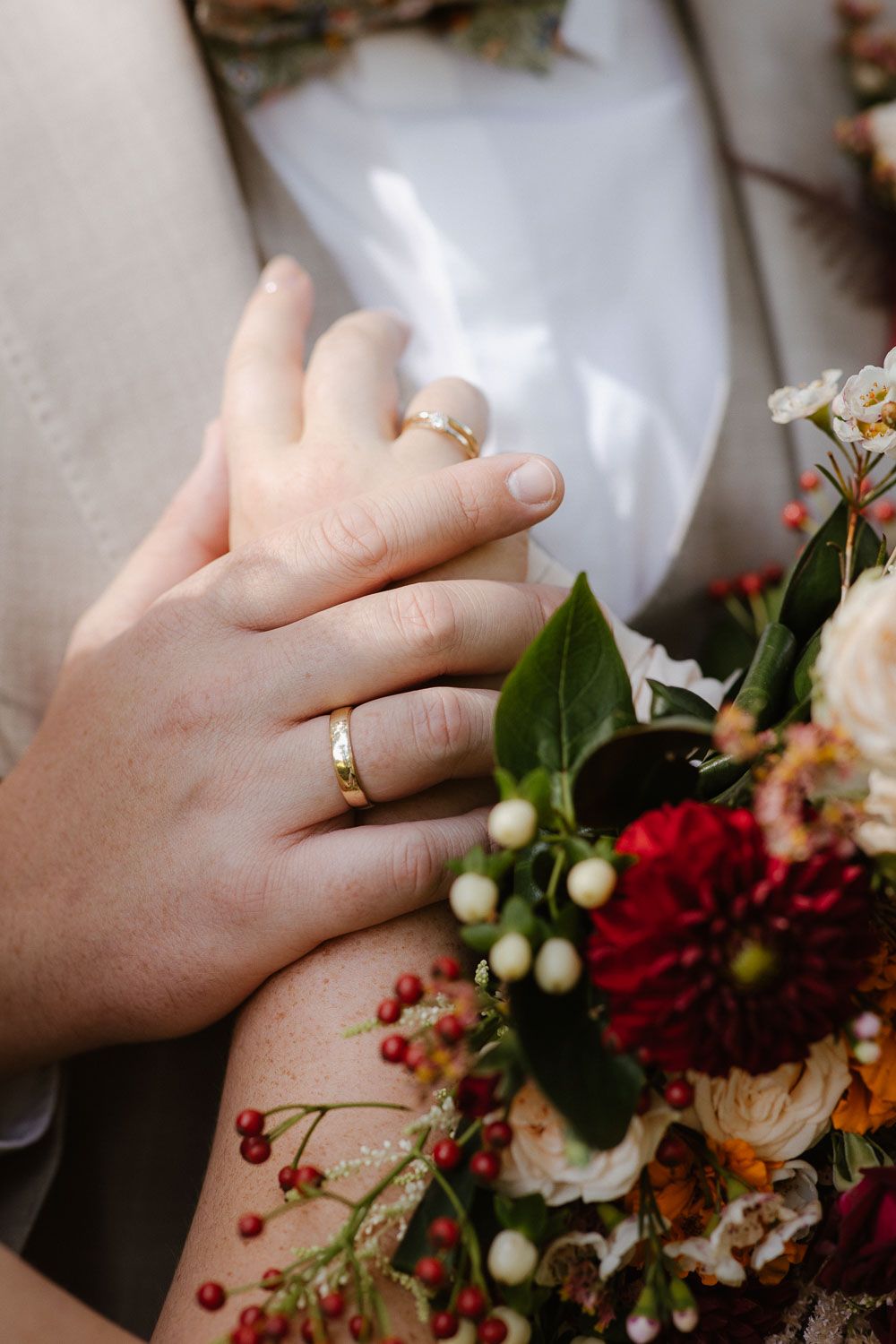 Close-up of newlywed couple hands with gold wedding rings resting on a colorful bridal bouquet.
