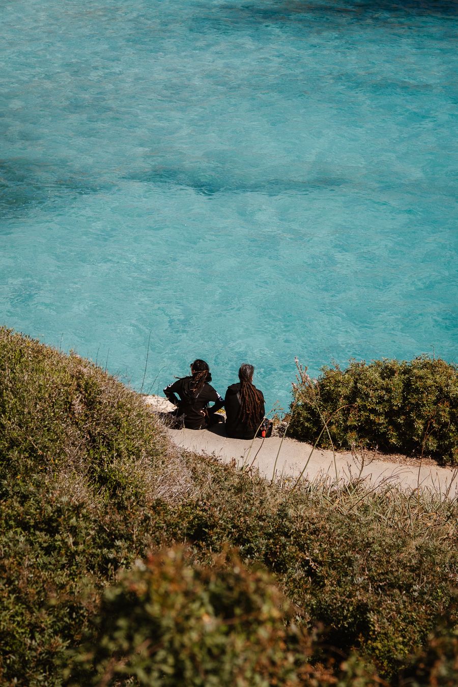 Two people sit on a rocky shoreline overlooking stunning turquoise blue ocean waters surrounded by green coastal shrubs.