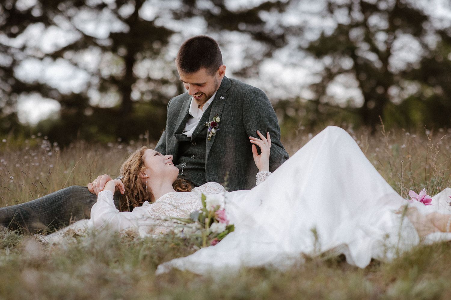 Romantic wedding couple lying in wildflower meadow, bride in white gown gazing lovingly at groom.