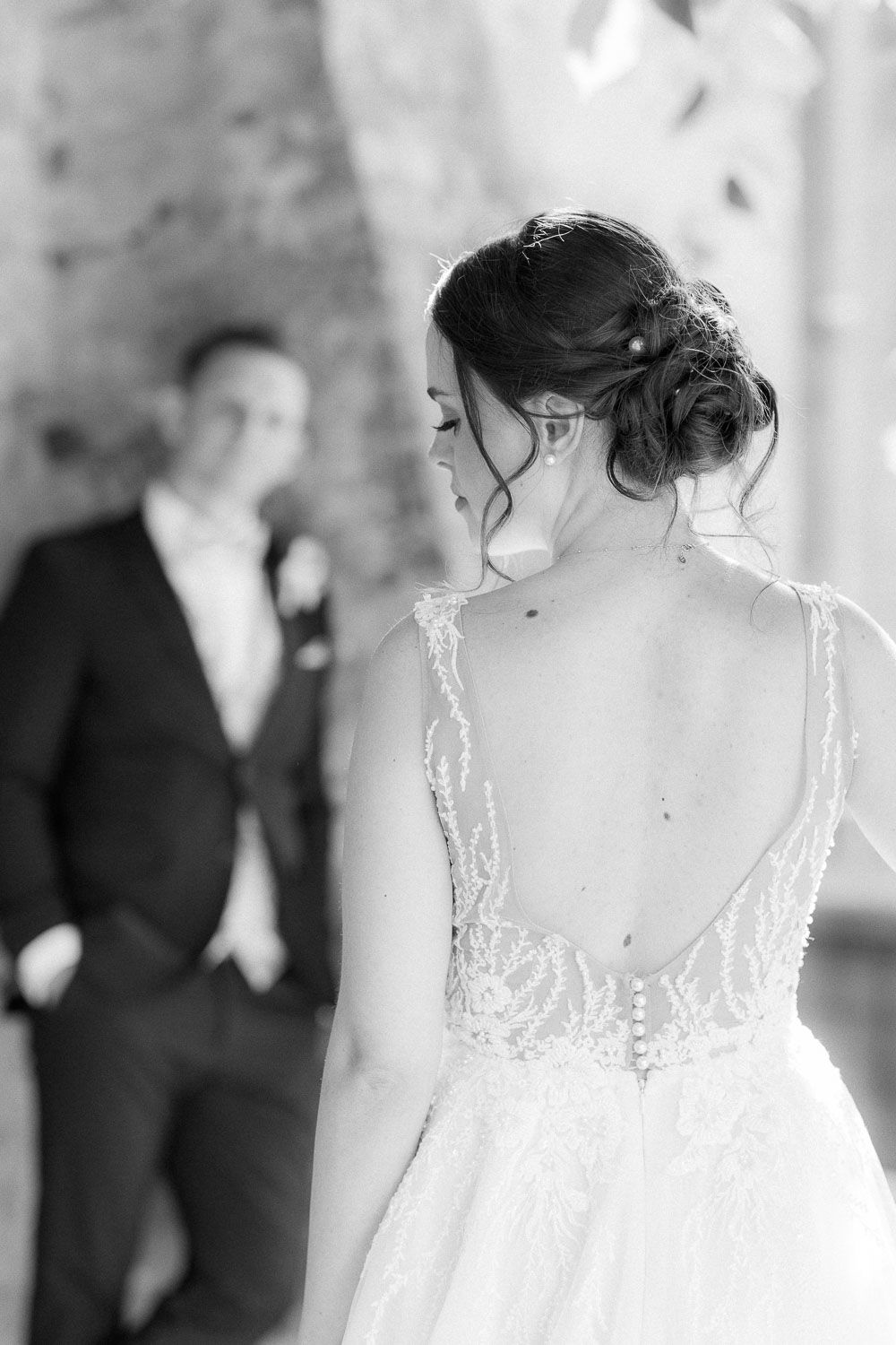 Black and white photo of bride in backless lace gown facing groom in suit during wedding ceremony.