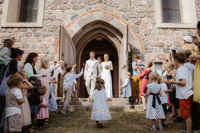 Newlyweds exiting a stone church through an arched doorway as guests and children line up to celebrate.