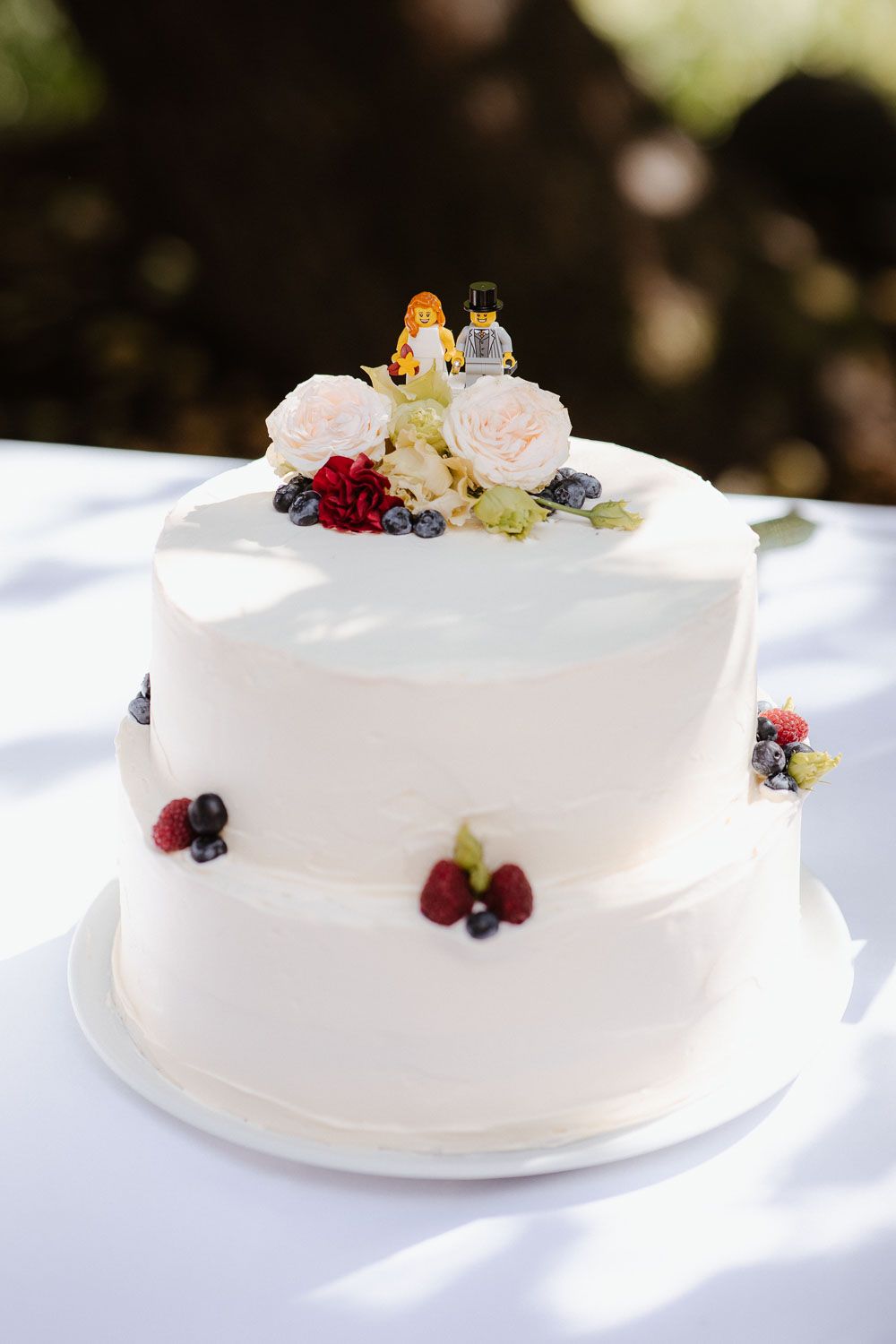 Elegant two-tier white wedding cake decorated with fresh berries, roses, and LEGO bride and groom toppers.
