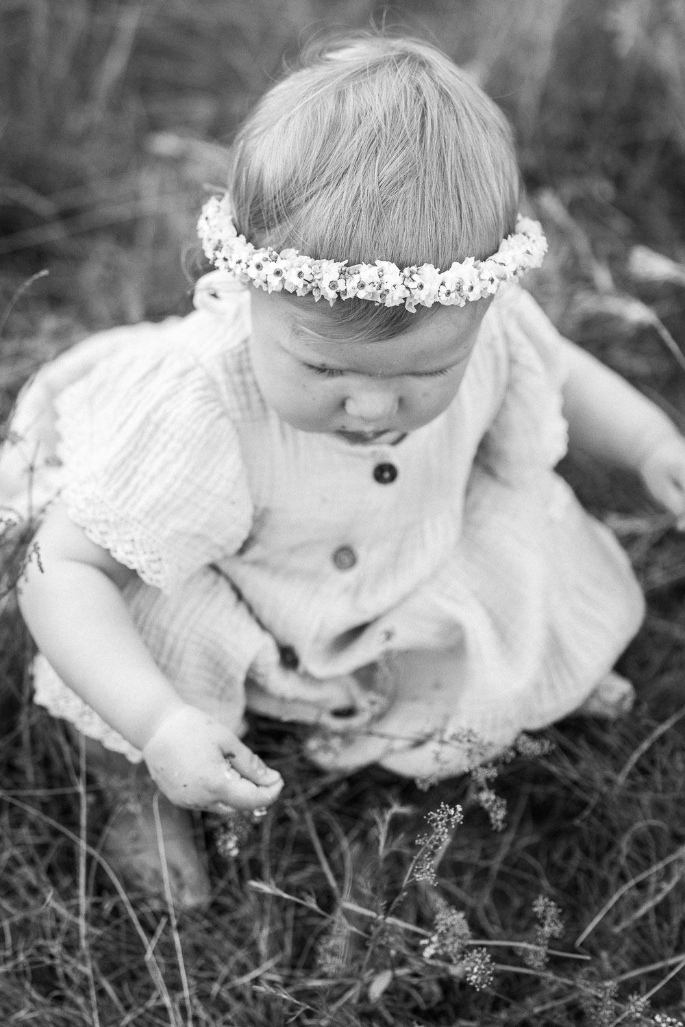 Black and white photo of baby girl in floral crown sitting in grass.