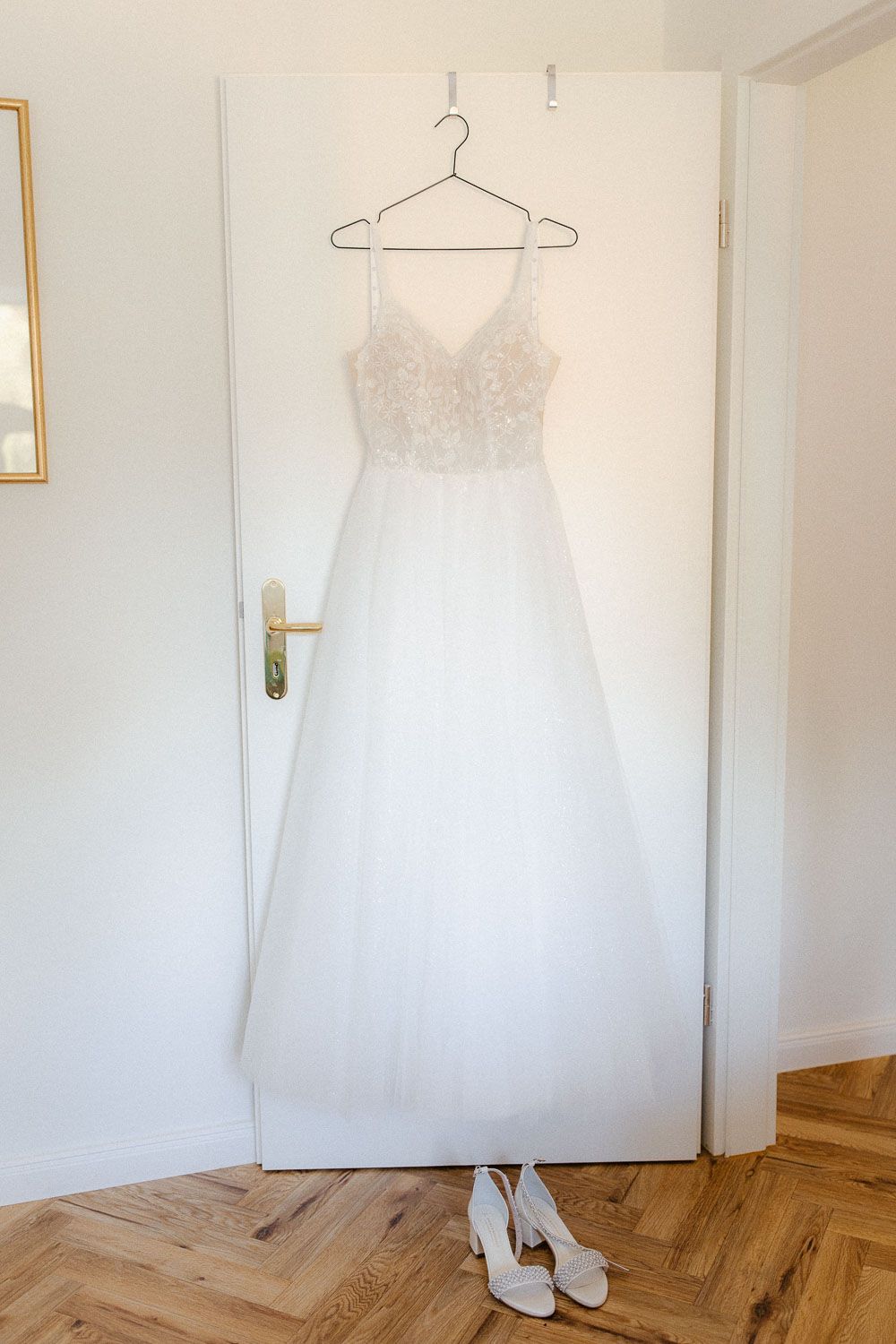 White lace A-line wedding dress hanging on door with matching bridal heels on hardwood floor below.