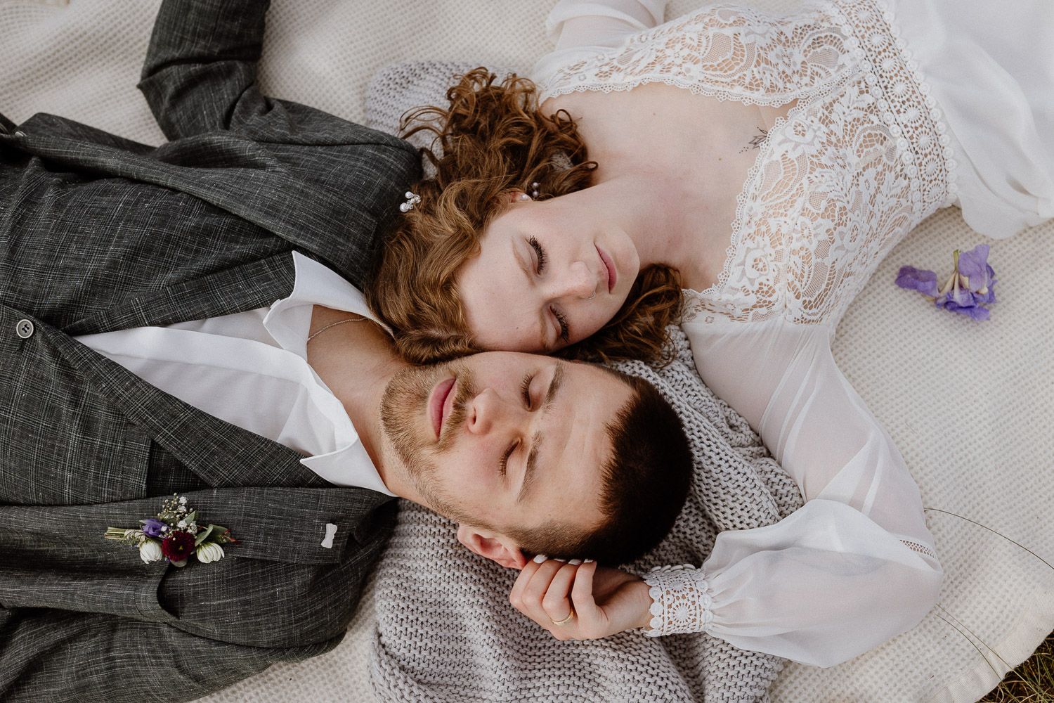 Overhead view of newlyweds lying together with eyes closed, bride in lace dress, groom in gray suit.