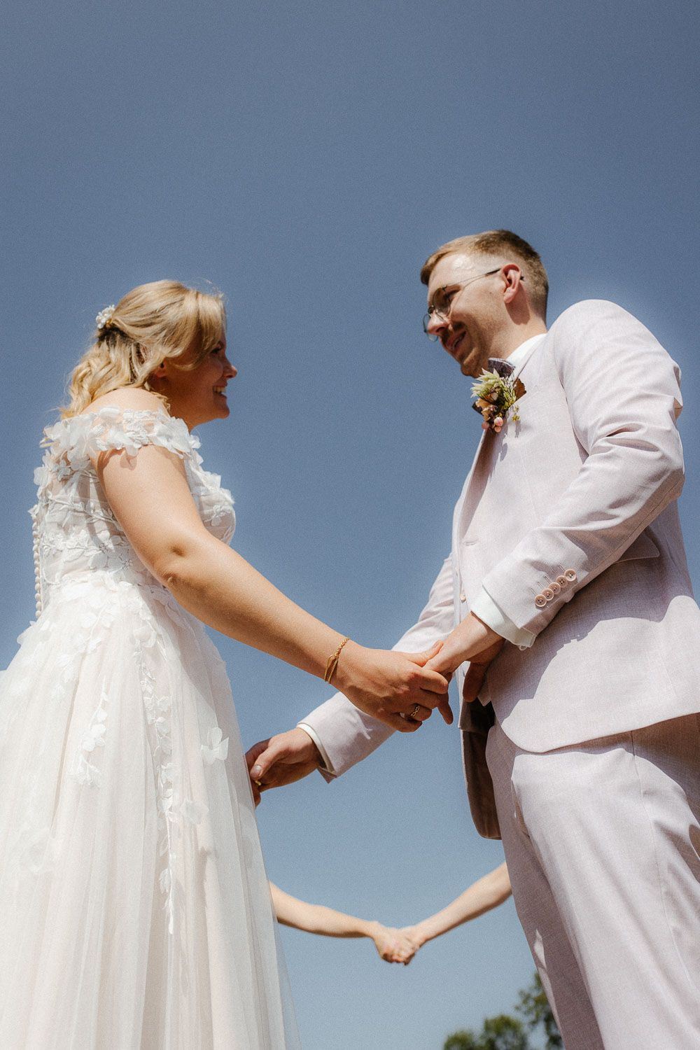 Bride and groom holding hands and smiling at each other against a clear blue sky on their wedding day.