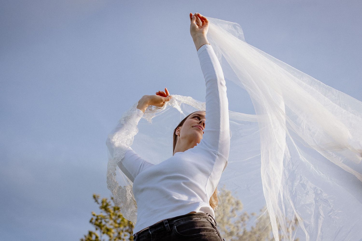 Woman in white top releasing flowing veil into blue sky outdoors.