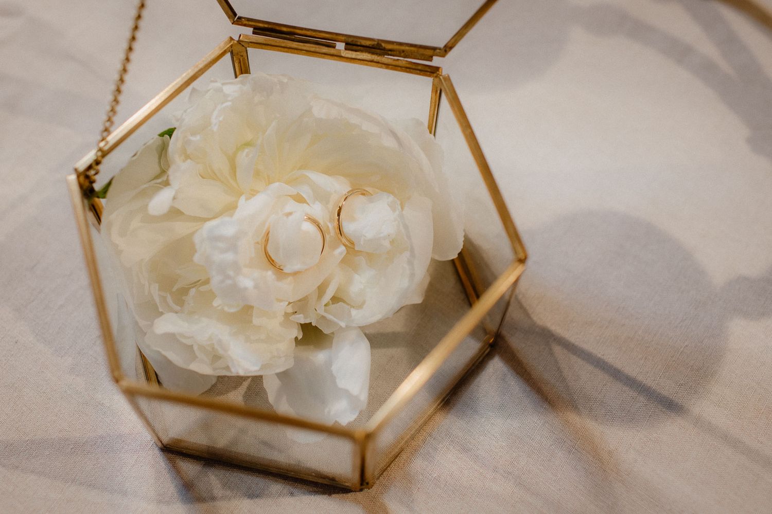 Wedding rings displayed in gold hexagonal glass terrarium with white peony flowers on marble surface.
