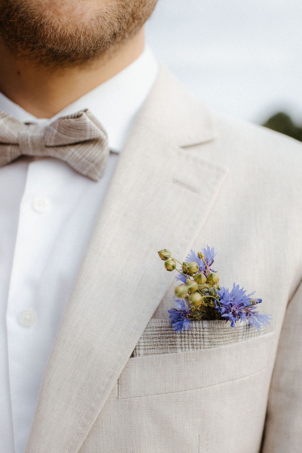 Groom in beige linen suit with bow tie and blue wildflower boutonniere in pocket at wedding.