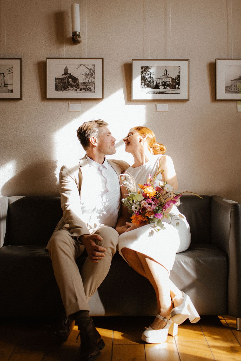 Happy newlywed couple sitting on a couch laughing together with a colorful bouquet in sunlit room.