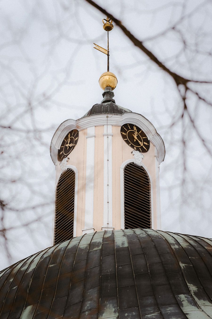 Historic church steeple with gold weather vane and ornate clock tower framed by bare winter branches.