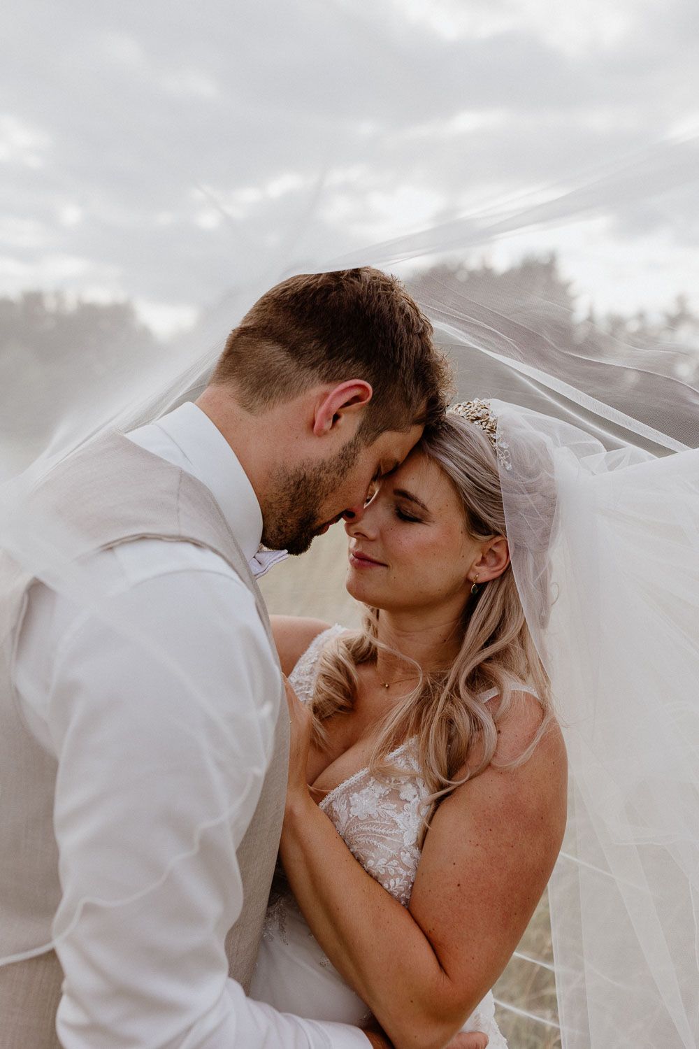 Bride and groom share intimate moment under flowing veil with cloudy sky backdrop at outdoor wedding.