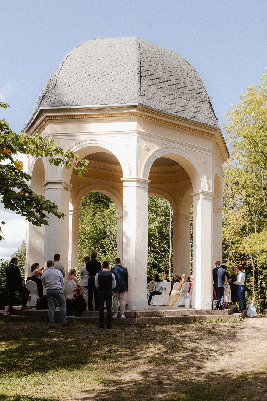 Wedding guests gathered under an elegant white domed pavilion surrounded by trees during an outdoor ceremony.