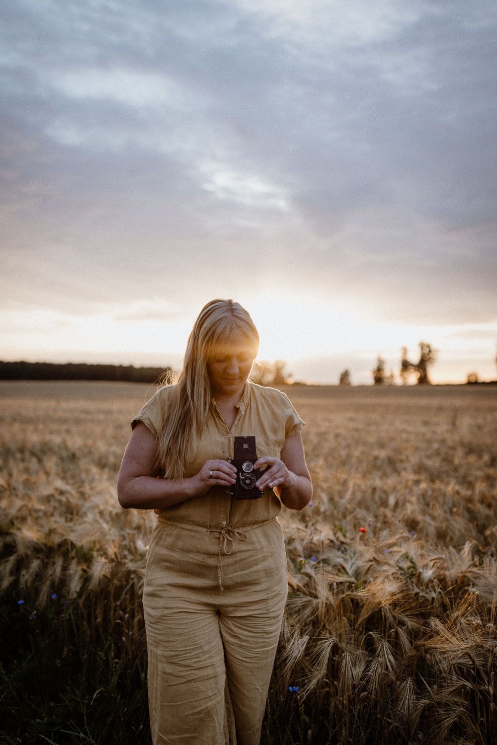 Woman in tan jumpsuit holding a vintage camera in a golden wheat field at sunset.
