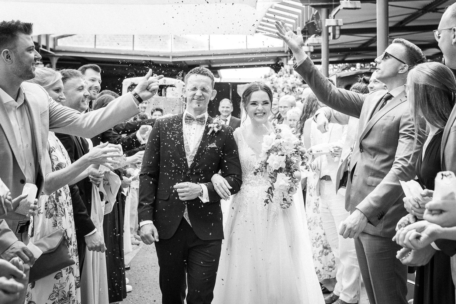 Black and white photo of newlyweds walking through confetti shower surrounded by cheering guests.