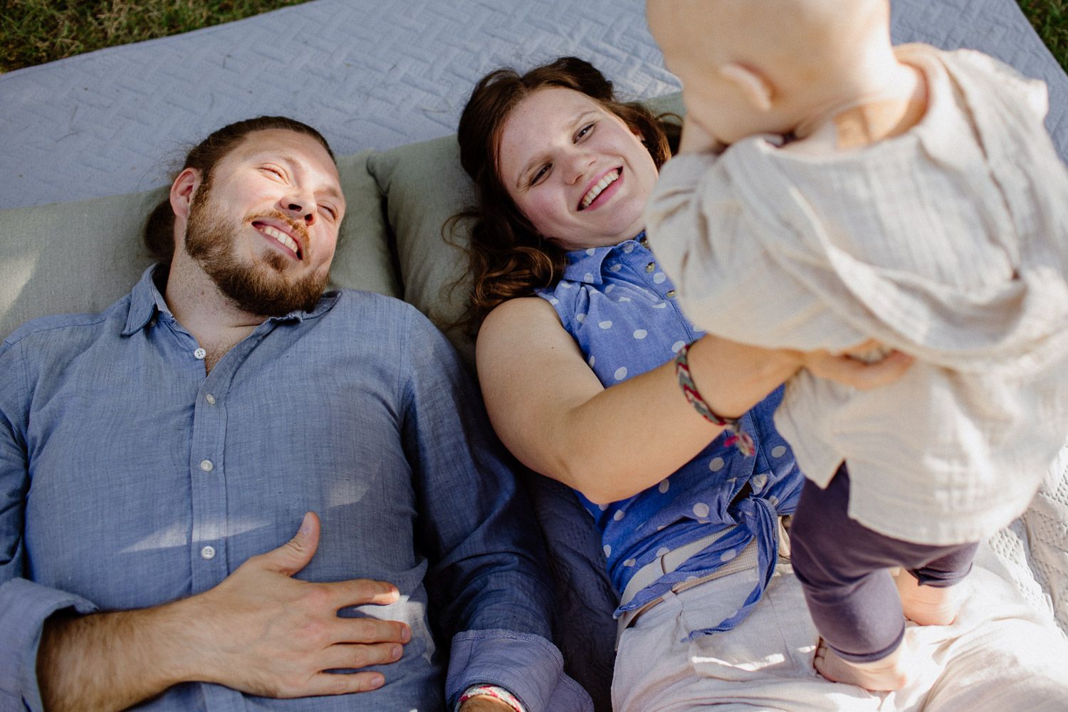 Happy family with parents lying on blanket smiling at standing toddler outdoors.