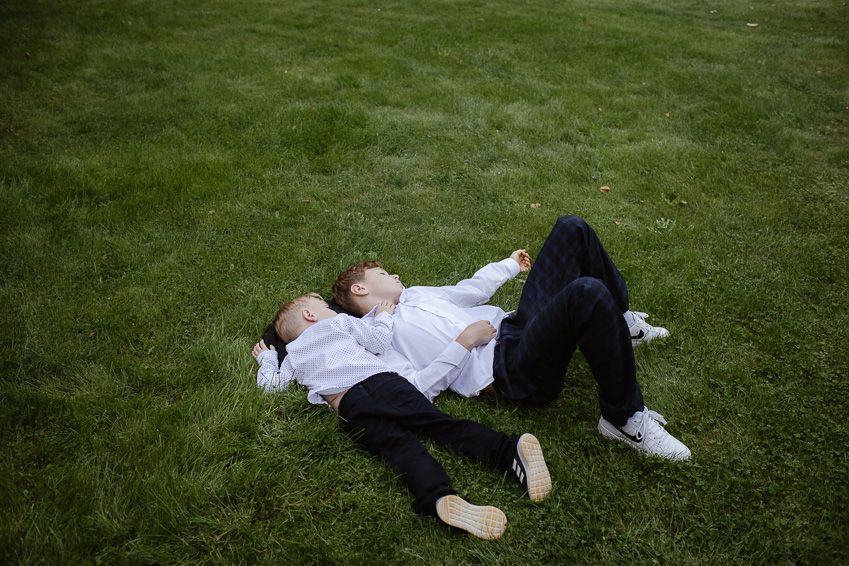 Two young boys in white shirts and black pants lying on green grass relaxing at a wedding reception.