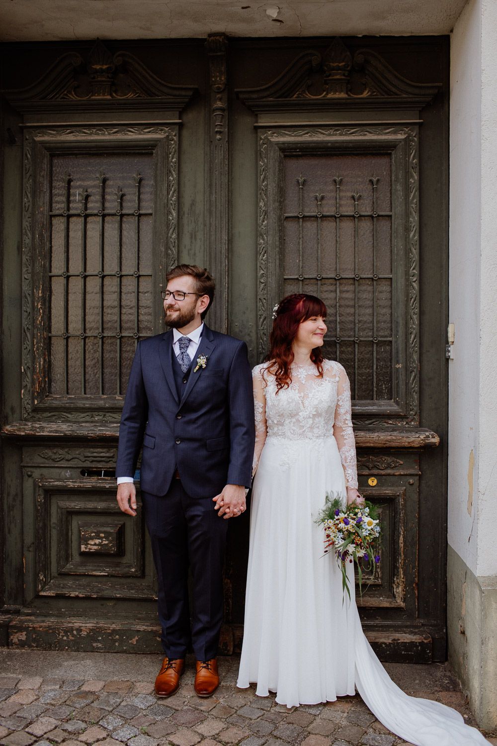 Bride and groom holding hands before ornate green vintage doors on cobblestone street.
