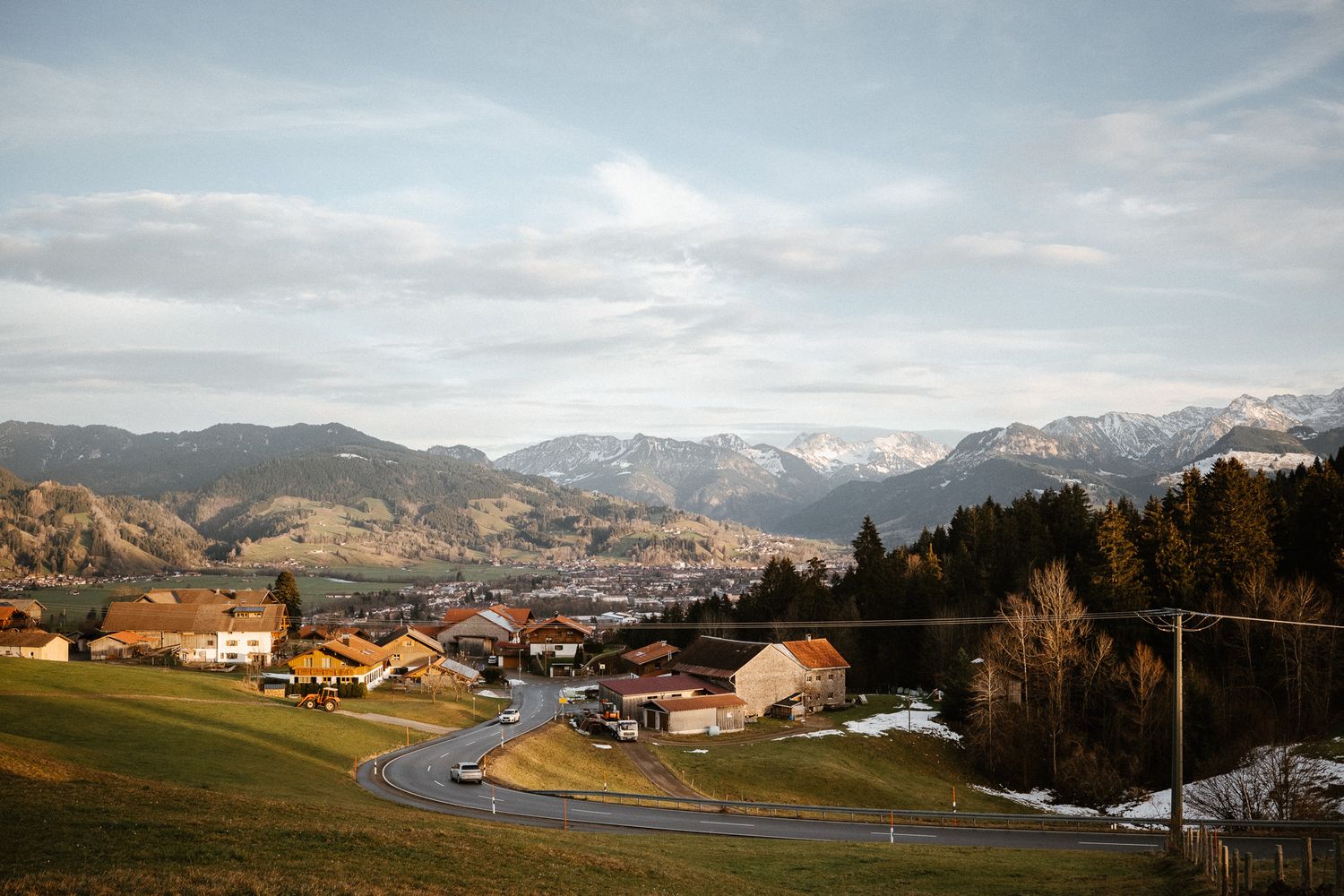 Scenic Alpine village with winding road, traditional chalets, and snow-capped mountain peaks at dusk.